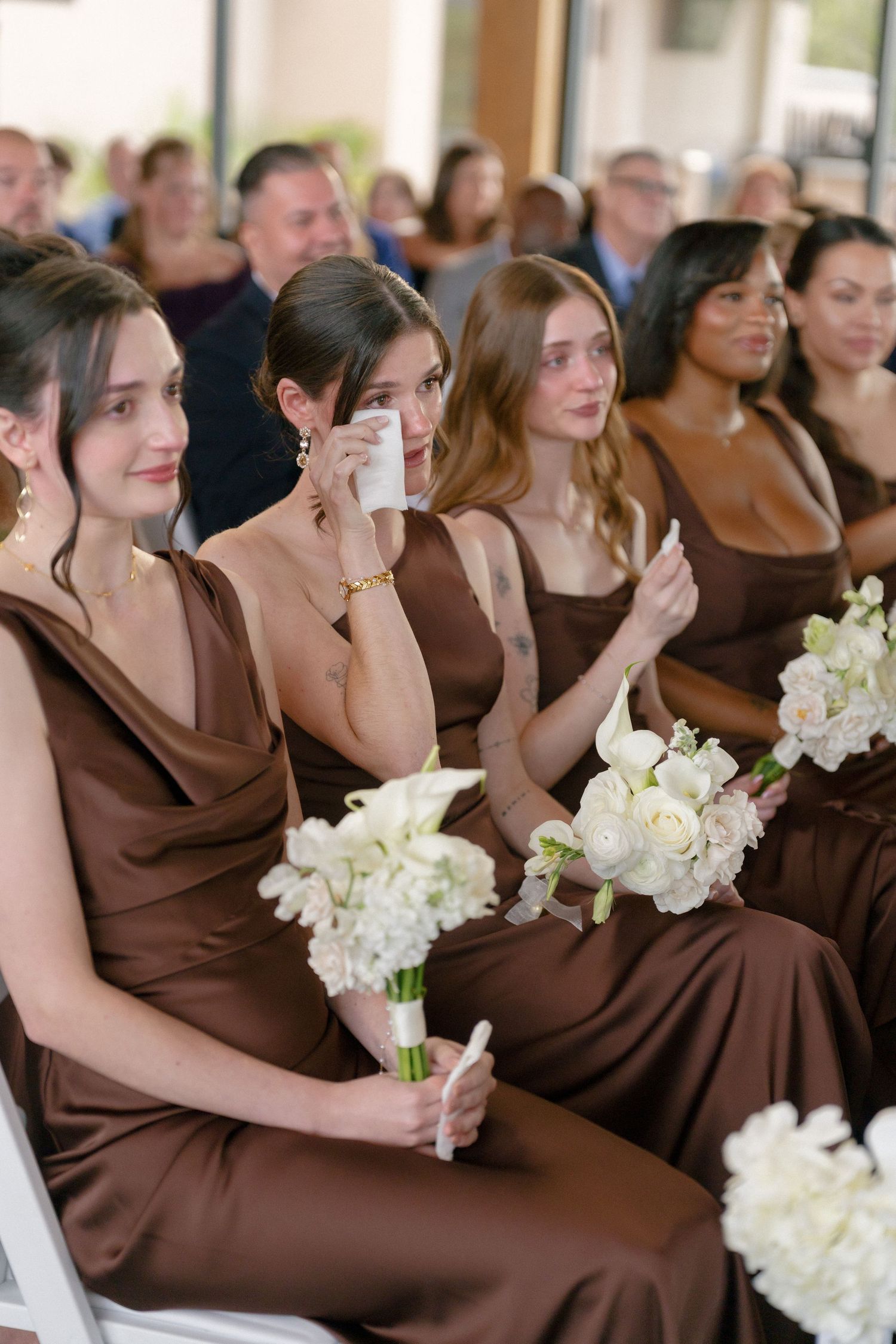 Emotional bridesmaids in brown dresses holding white bouquets during wedding ceremony.