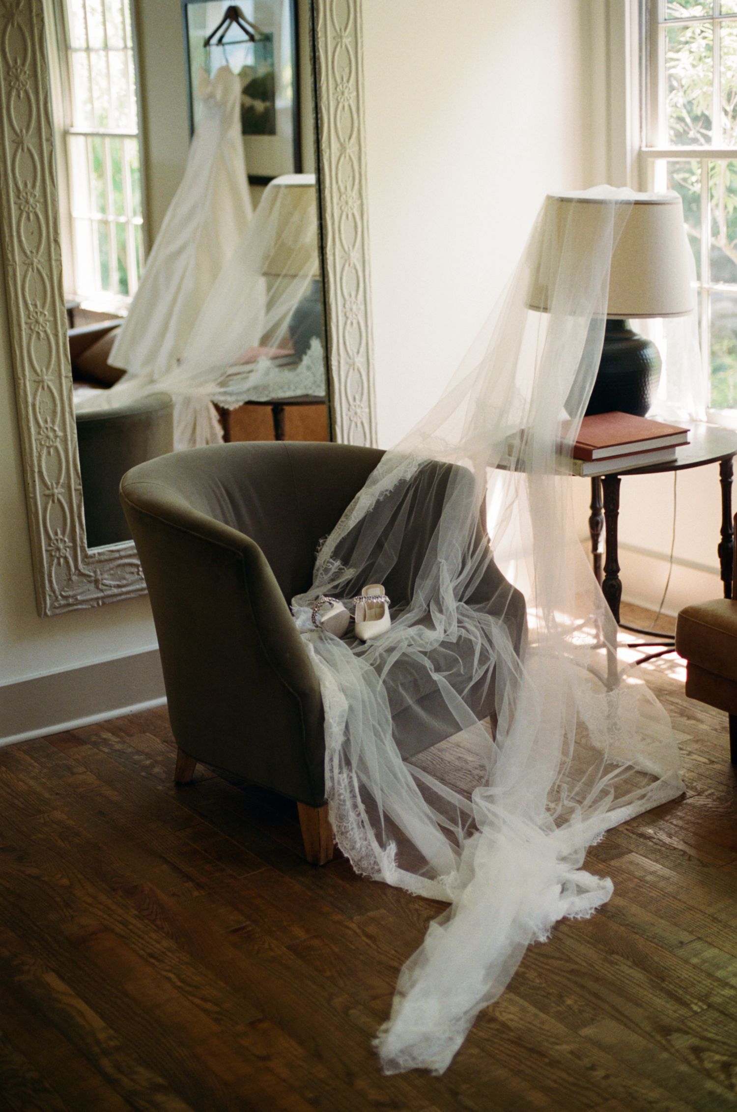 Bridal veil draped over gray armchair with wedding dress reflected in ornate mirror, elegant wedding preparation scene.
