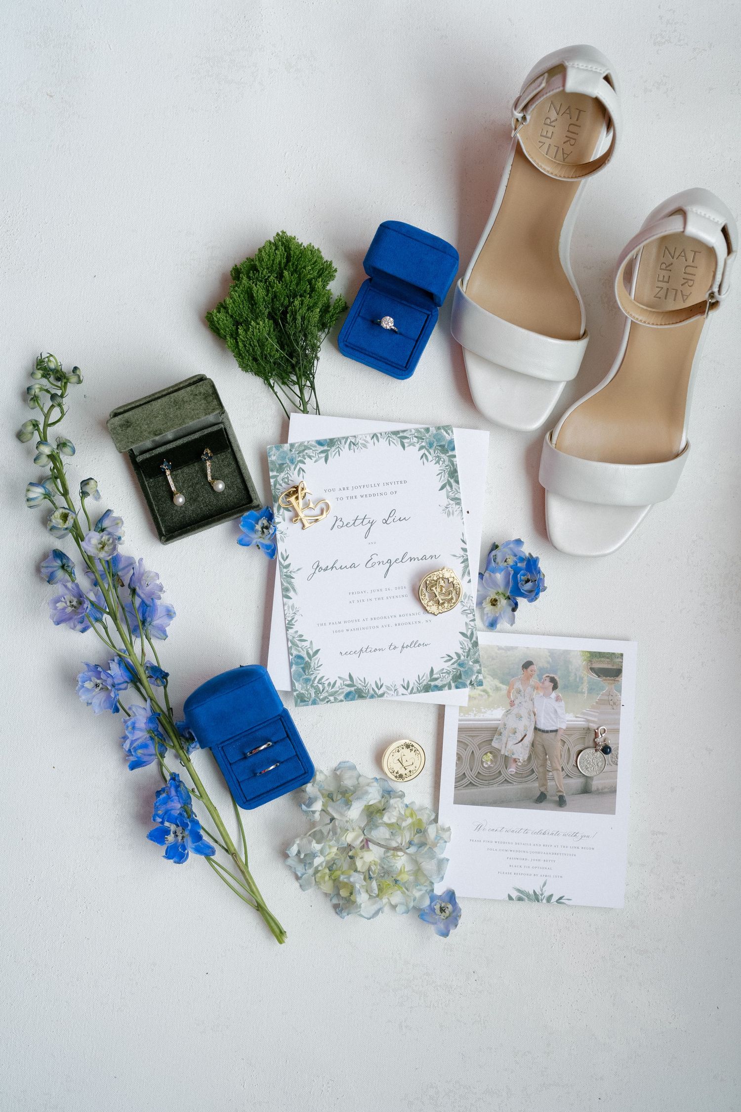 Elegant wedding flat lay featuring white heels, blue velvet ring boxes, invitations, and floral details.
