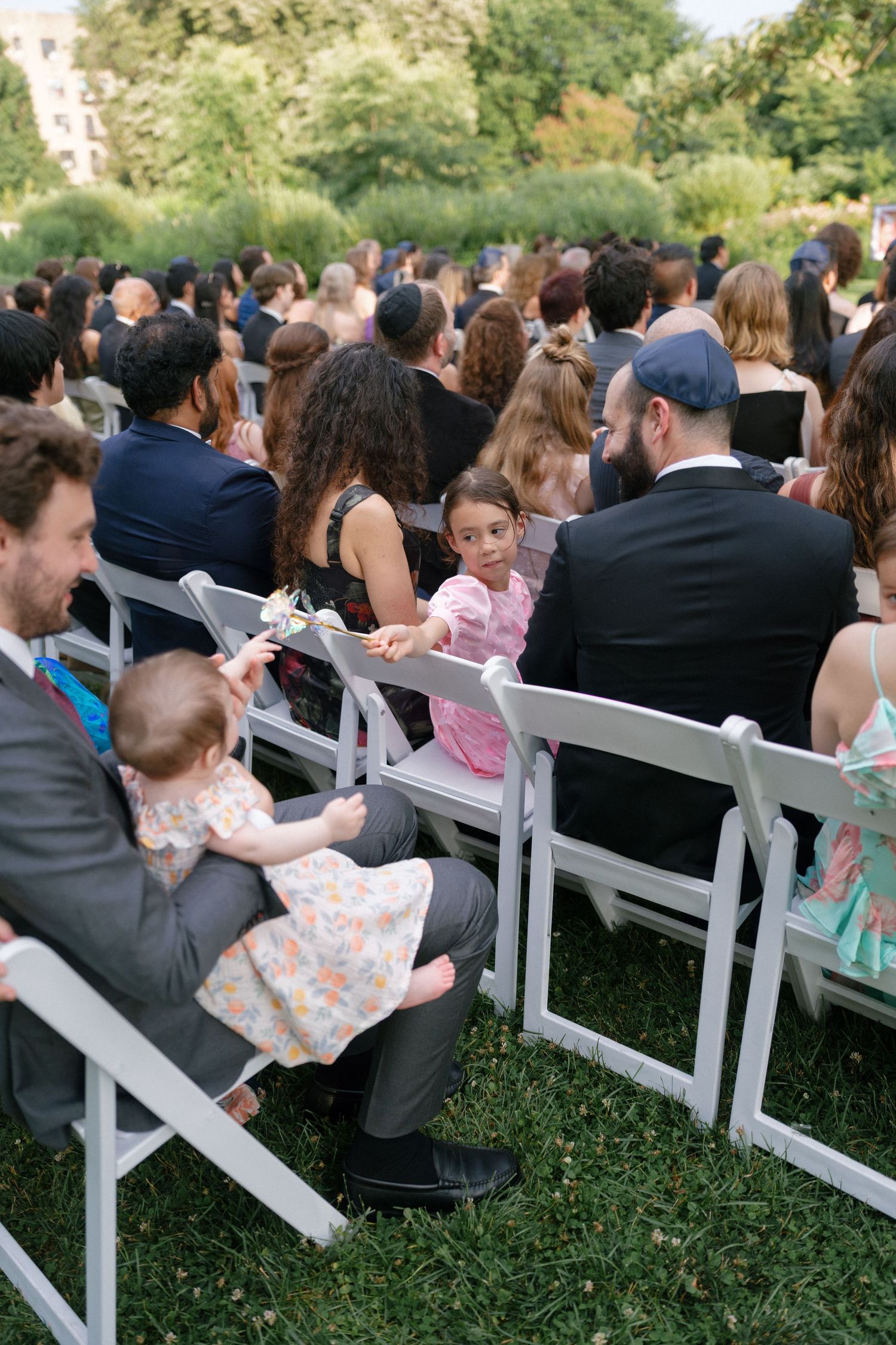 Wedding guests seated in white chairs at an outdoor garden ceremony with children in attendance.