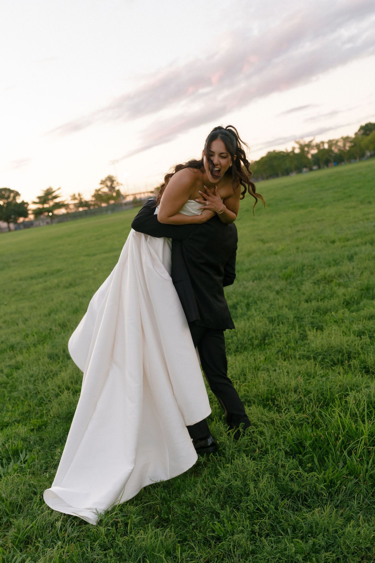 Joyful groom lifting laughing bride in white wedding gown on green field during golden hour sunset celebration.