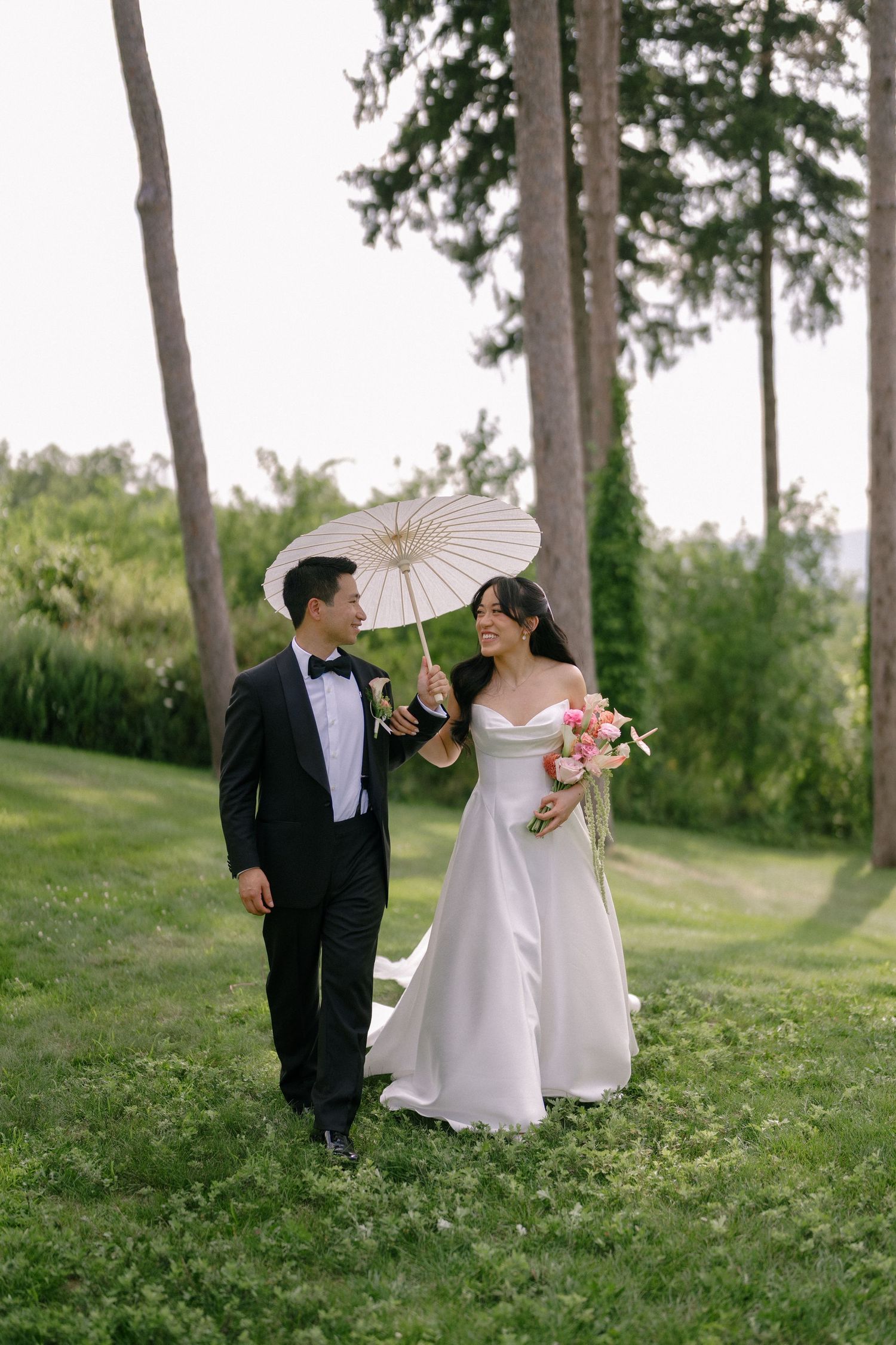 Smiling bride and groom walk through a green garden, sharing a white parasol while bride holds a pink bouquet.