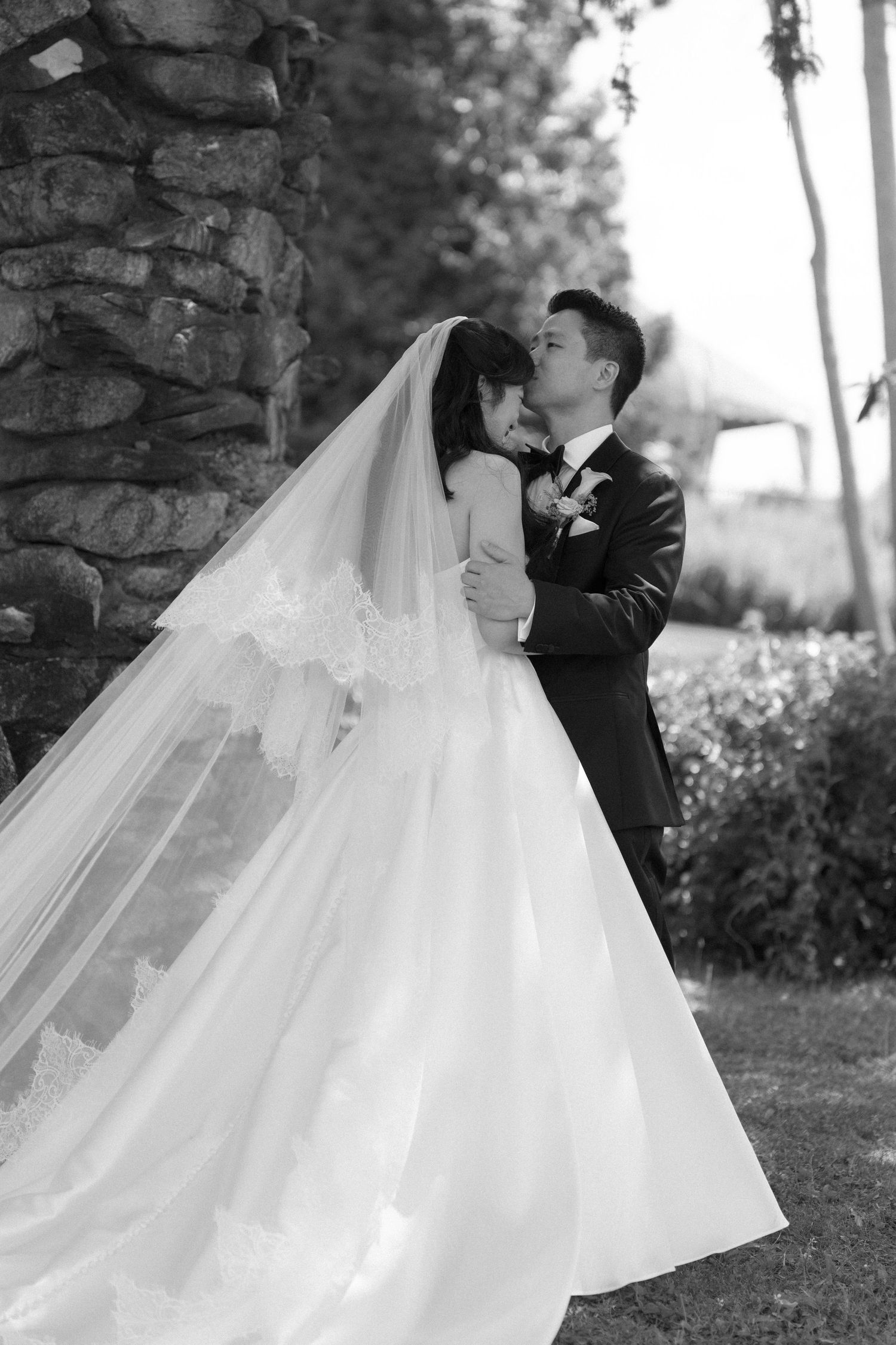 Black and white photo of a bride and groom embracing outdoors near a stone wall, her veil flowing gracefully.