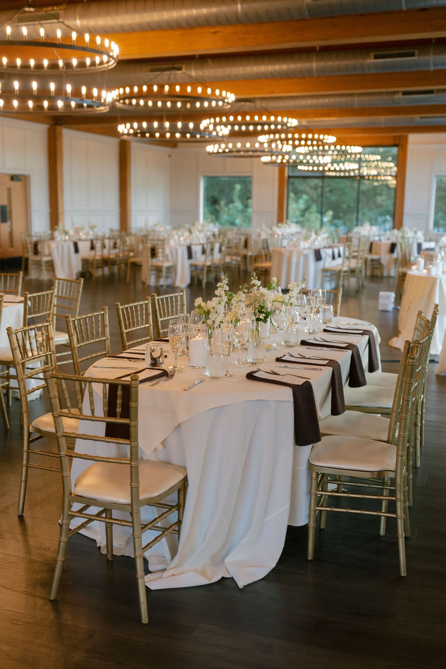 Elegant wedding reception hall with round tables, gold chiavari chairs, and circular chandeliers.