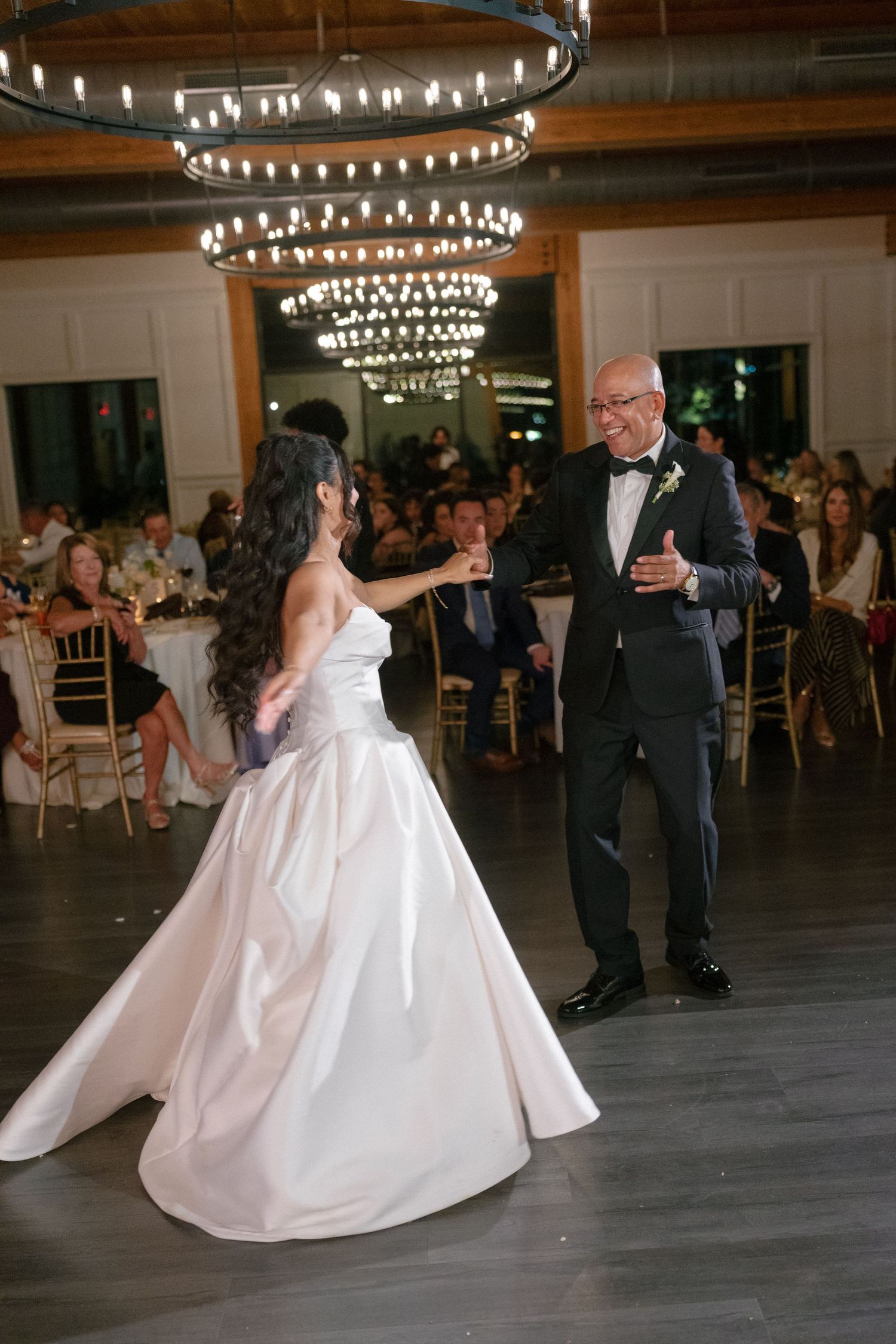 Bride and older gentleman share a dance at wedding reception under sparkling chandeliers.