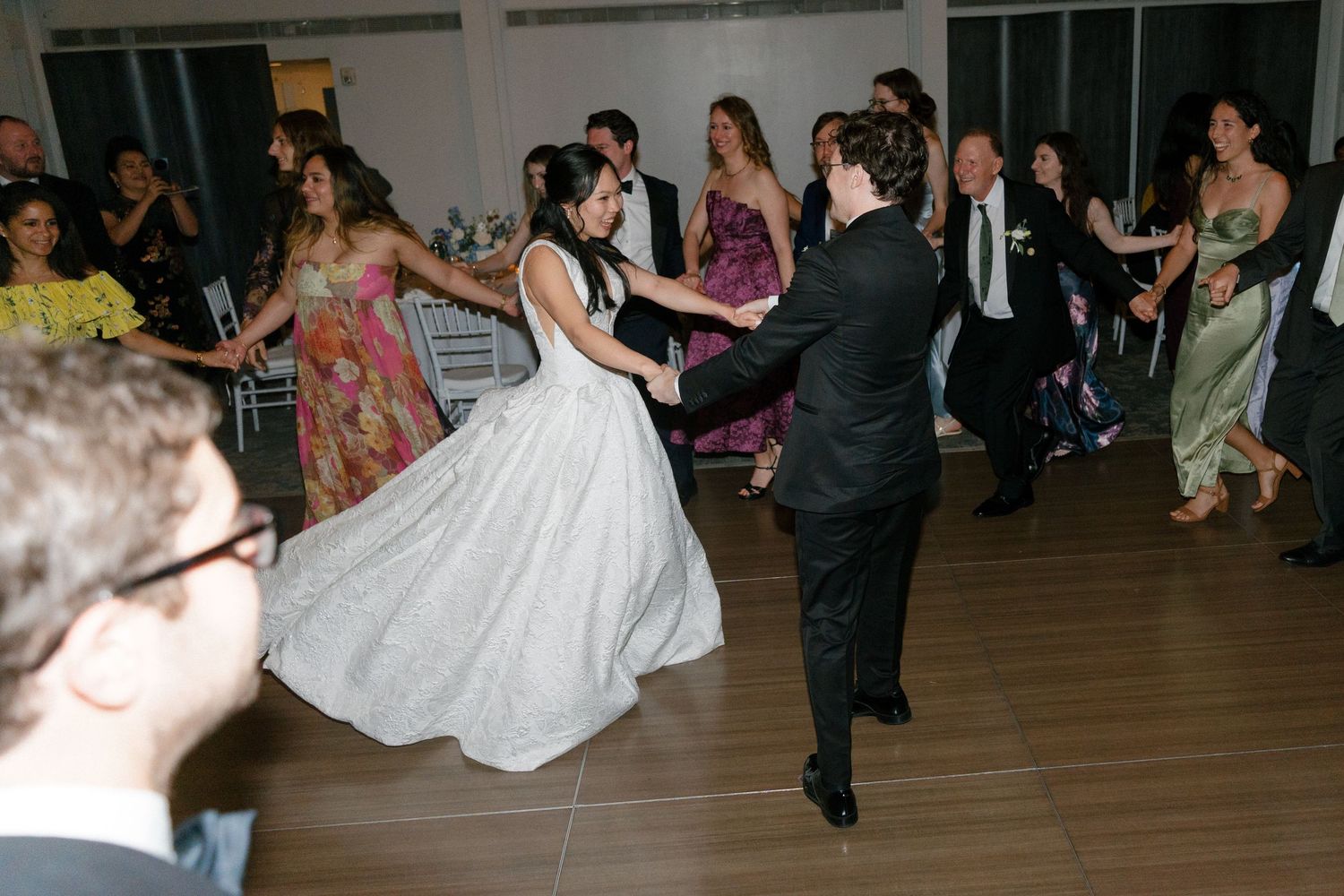 Bride and groom dance joyfully surrounded by guests at a lively indoor wedding reception celebration.