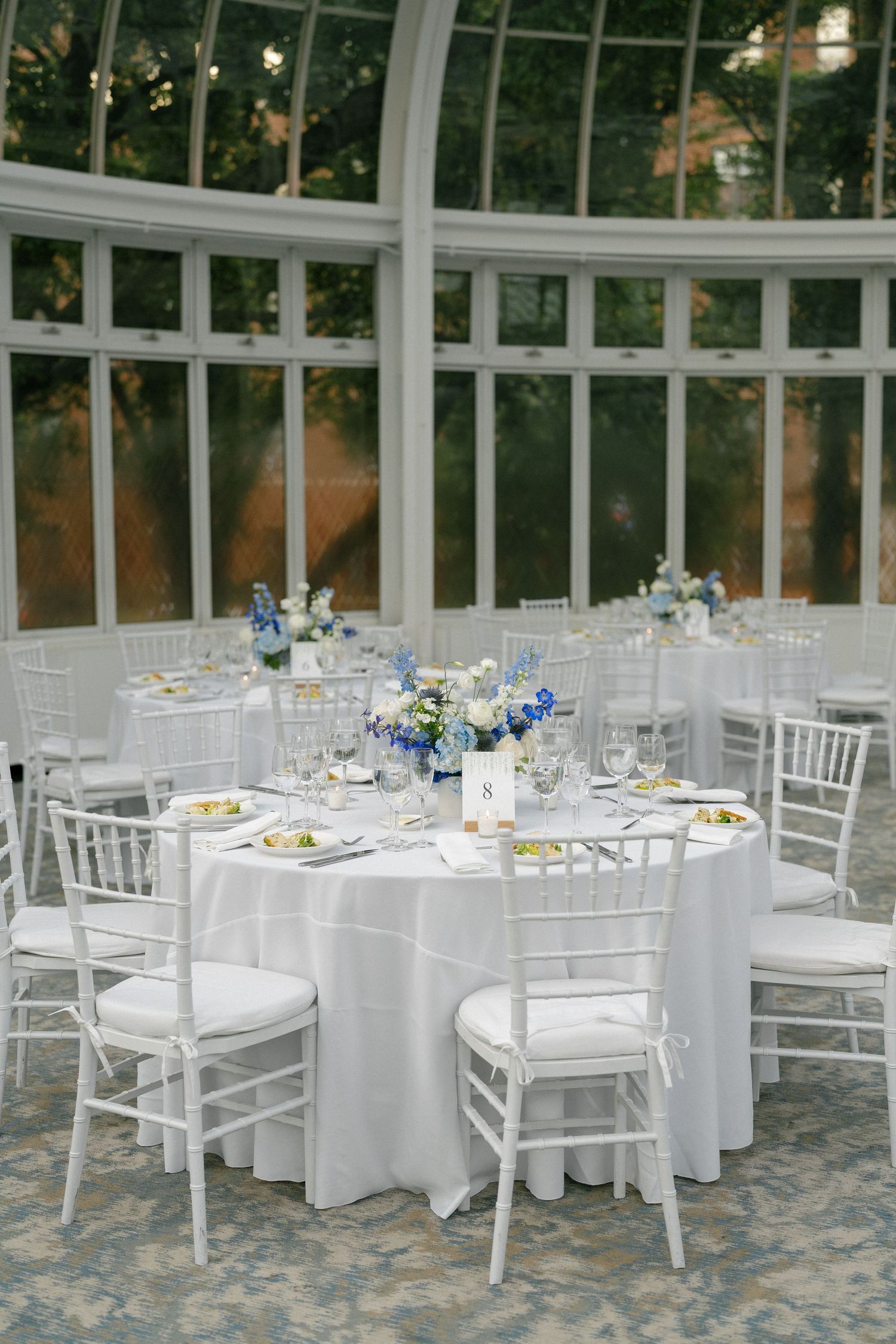 Elegant wedding reception tables with white linens, chiavari chairs, and blue floral centerpieces in a greenhouse venue.