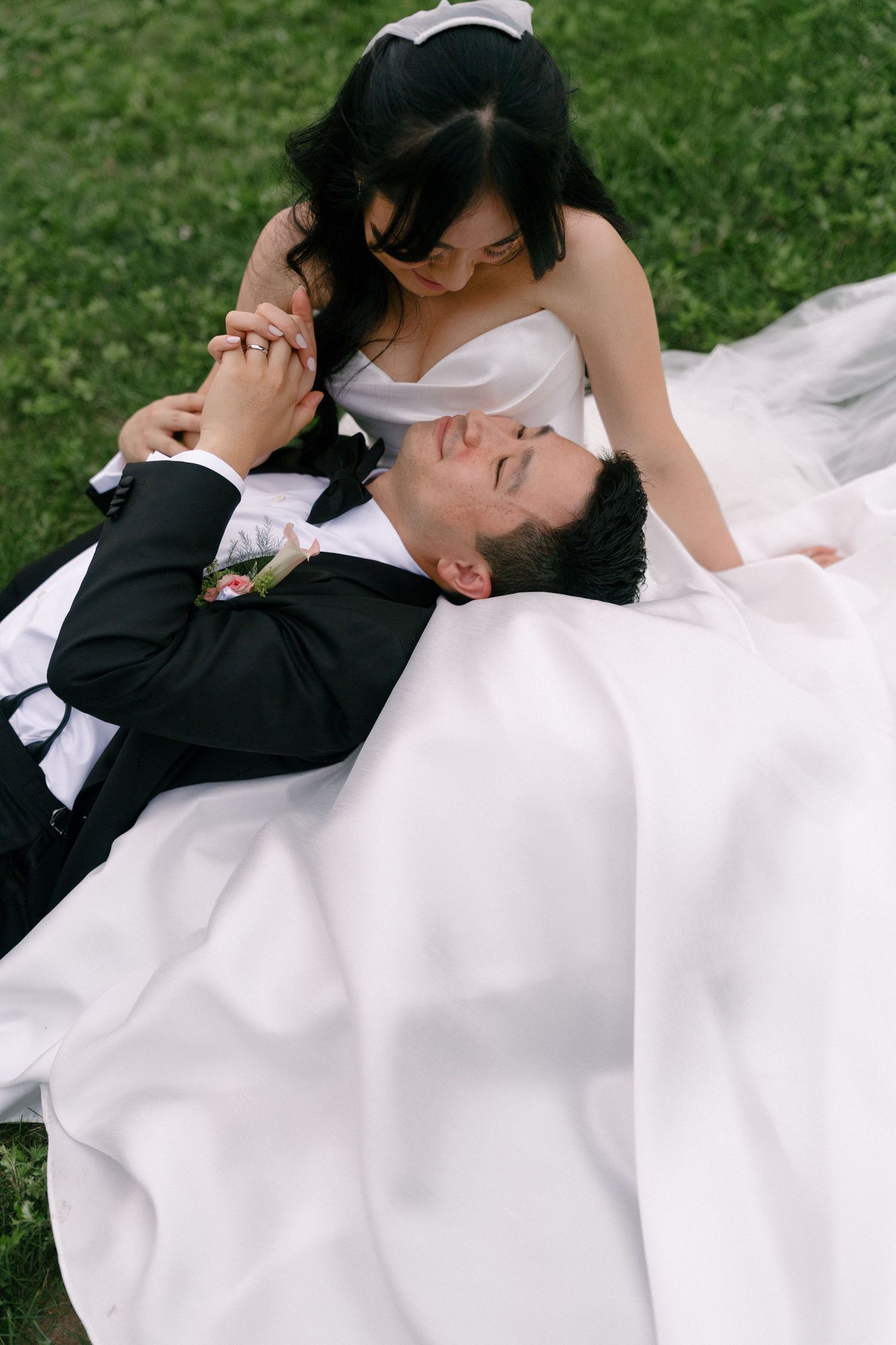 Groom lying on grass smiling up at bride in flowing white gown during outdoor portrait session.