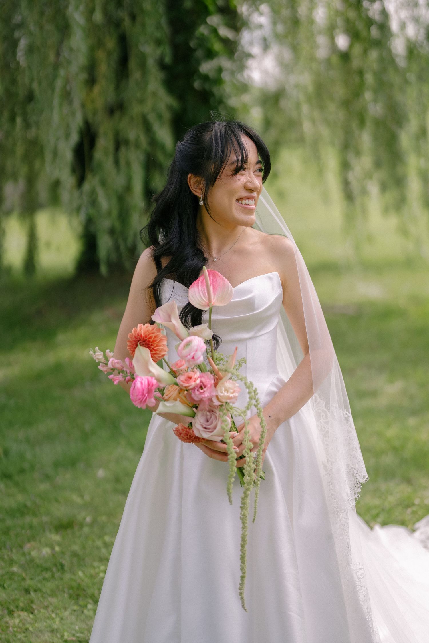 Smiling bride in strapless white gown holds colorful cascading bouquet before weeping willow tree.