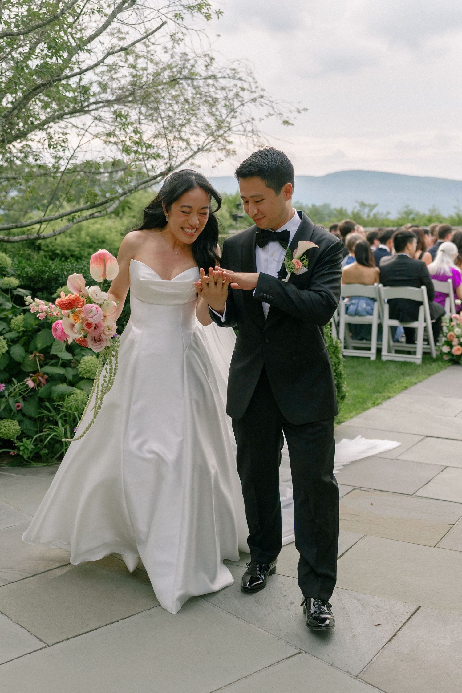 Newlyweds admiring wedding rings after outdoor ceremony with mountain views, bride holding pink bouquet in white gown.