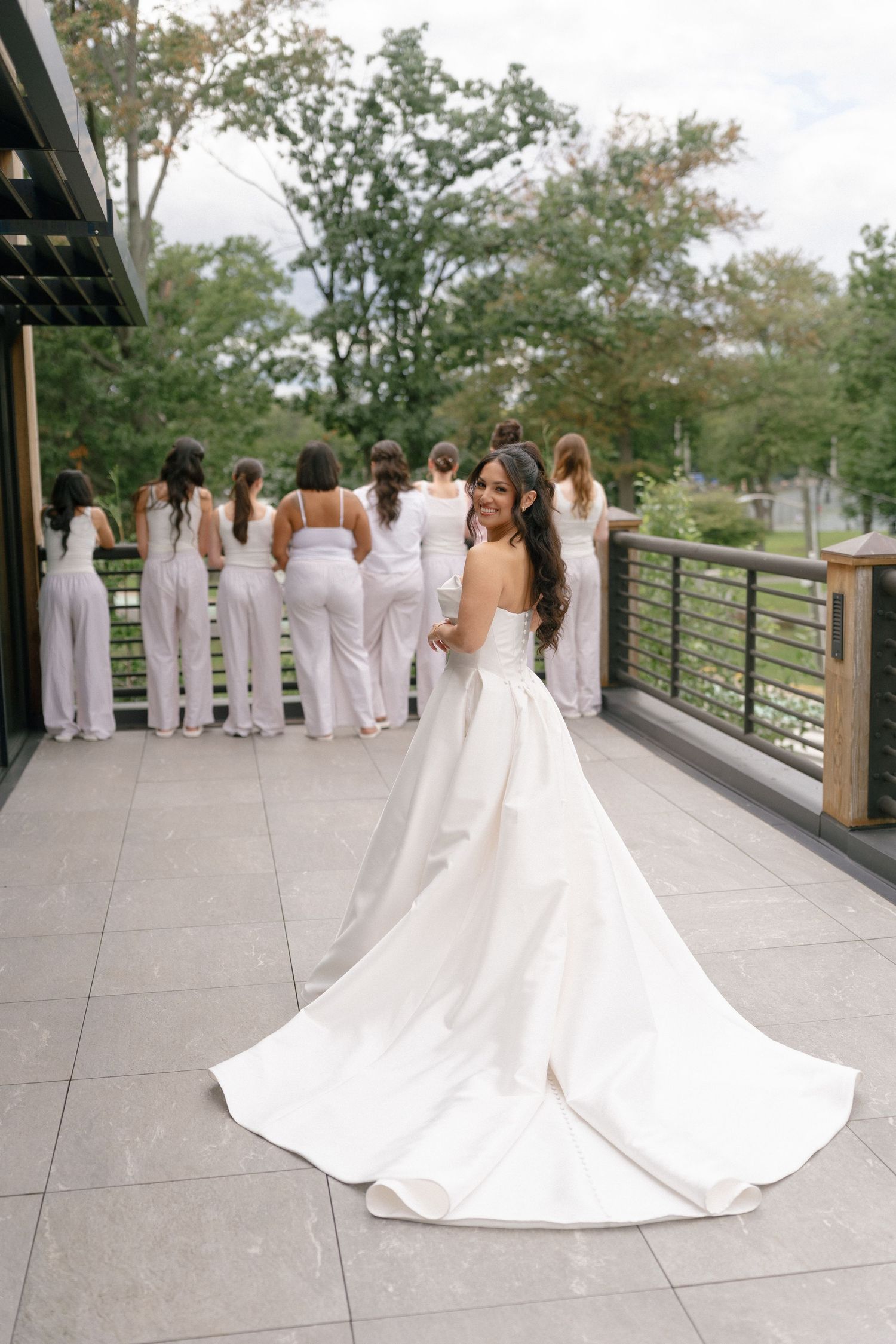 Bride in ball gown with long train turns to smile while bridesmaids in white stand on terrace.