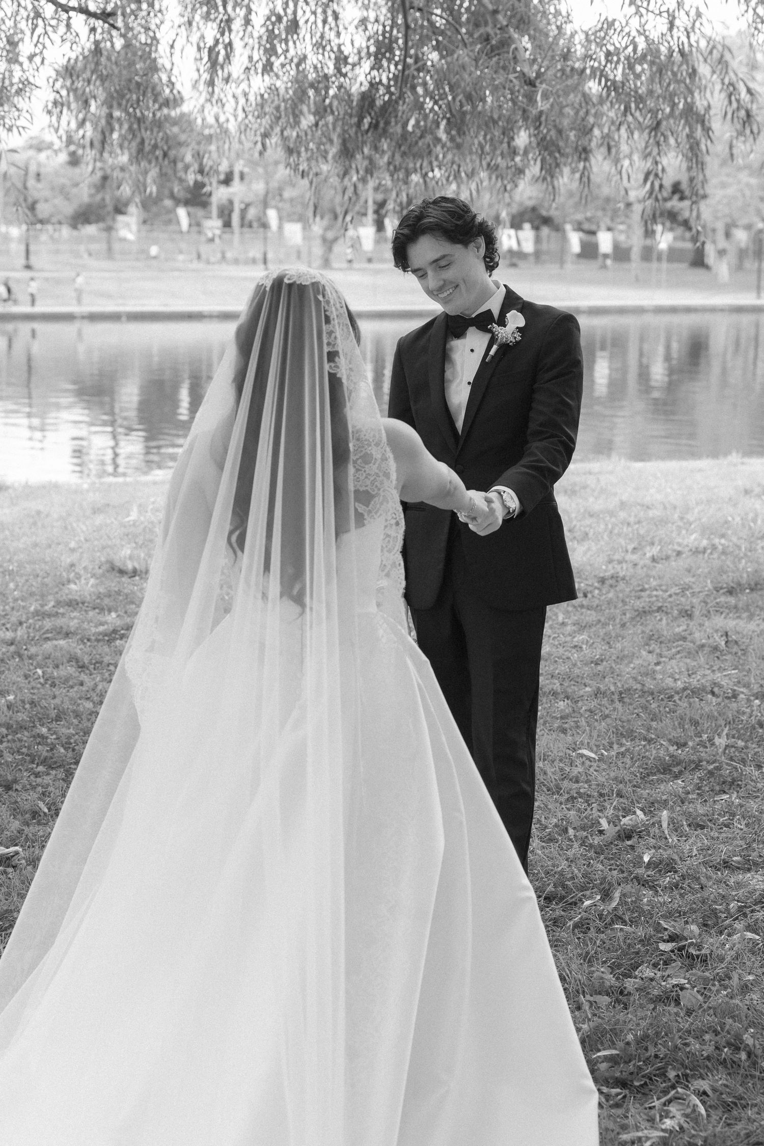 Groom smiles at veiled bride while holding hands near a serene pond in black and white.