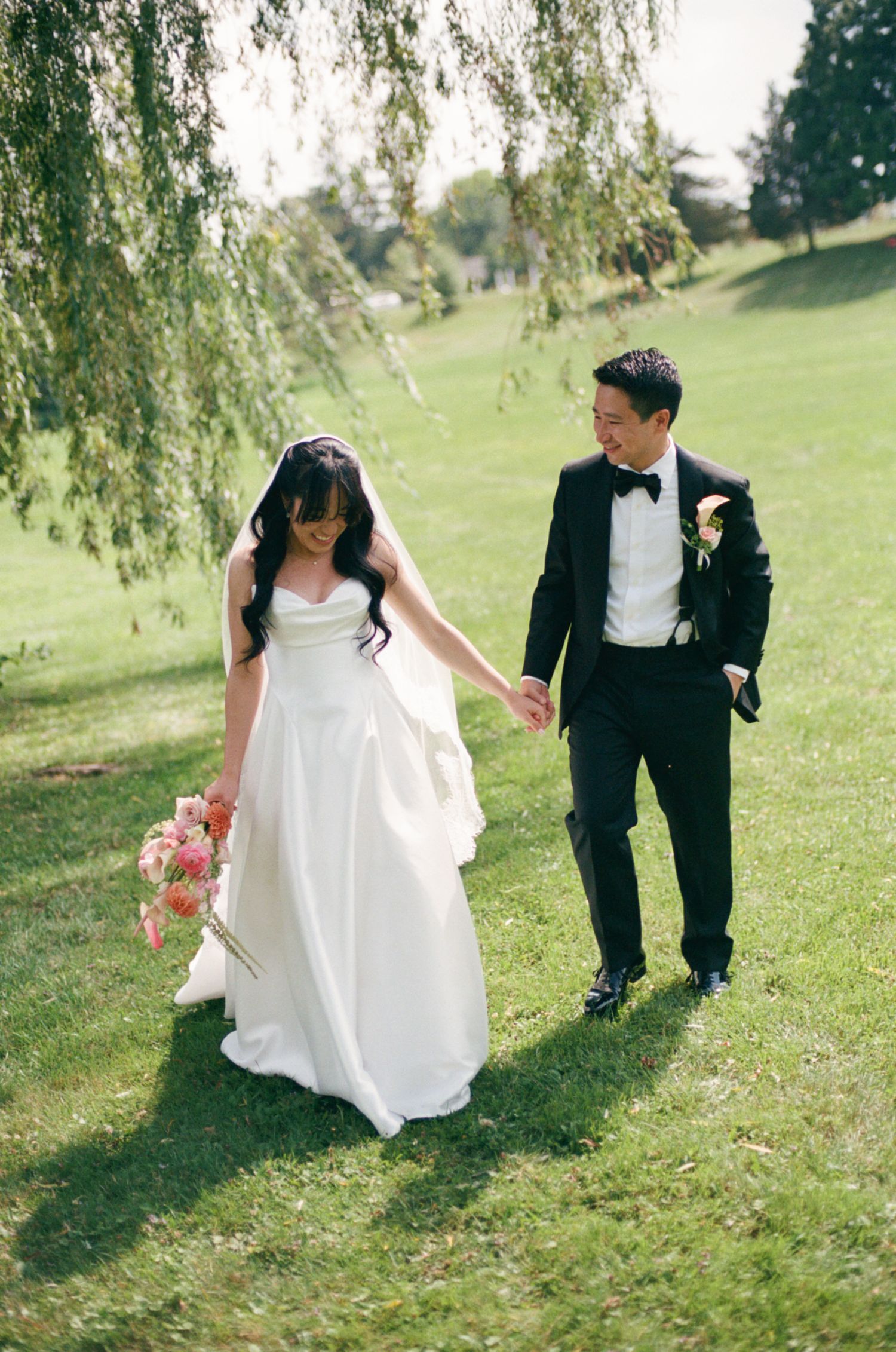 Bride in white gown and groom in black tuxedo holding hands walking on green grass under willow tree.