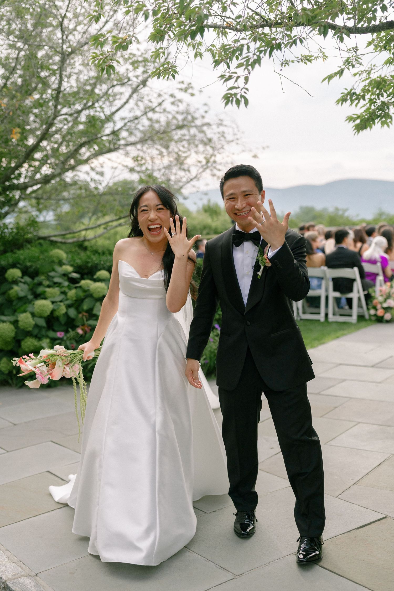Newlyweds joyfully show wedding rings after outdoor ceremony with mountain views and floral decor.