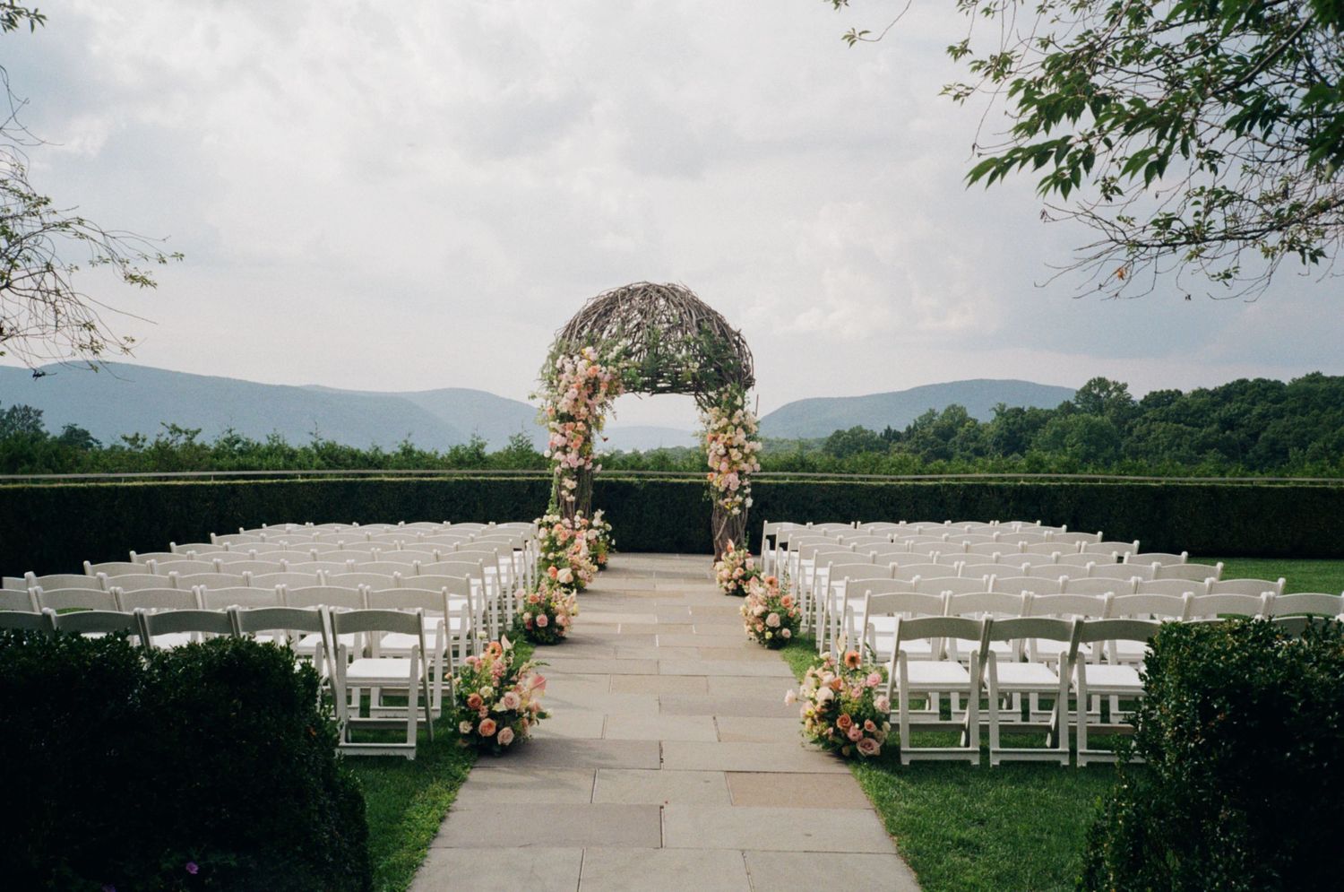 Outdoor wedding ceremony setup with white chairs, floral arch, and mountain views under a cloudy sky.
