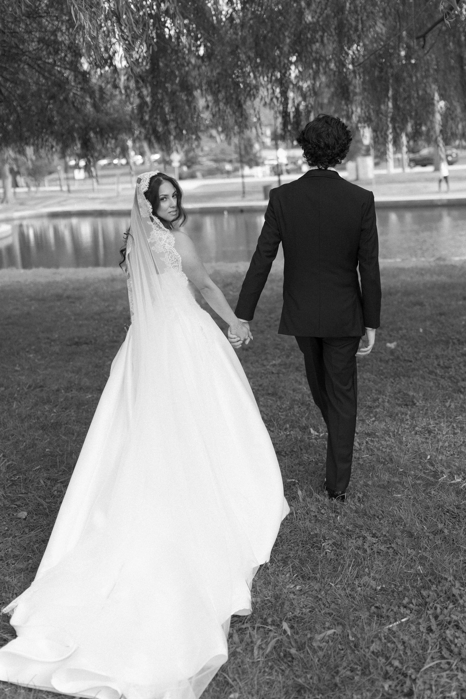Black and white photo of bride and groom walking hand in hand through a park near a pond.