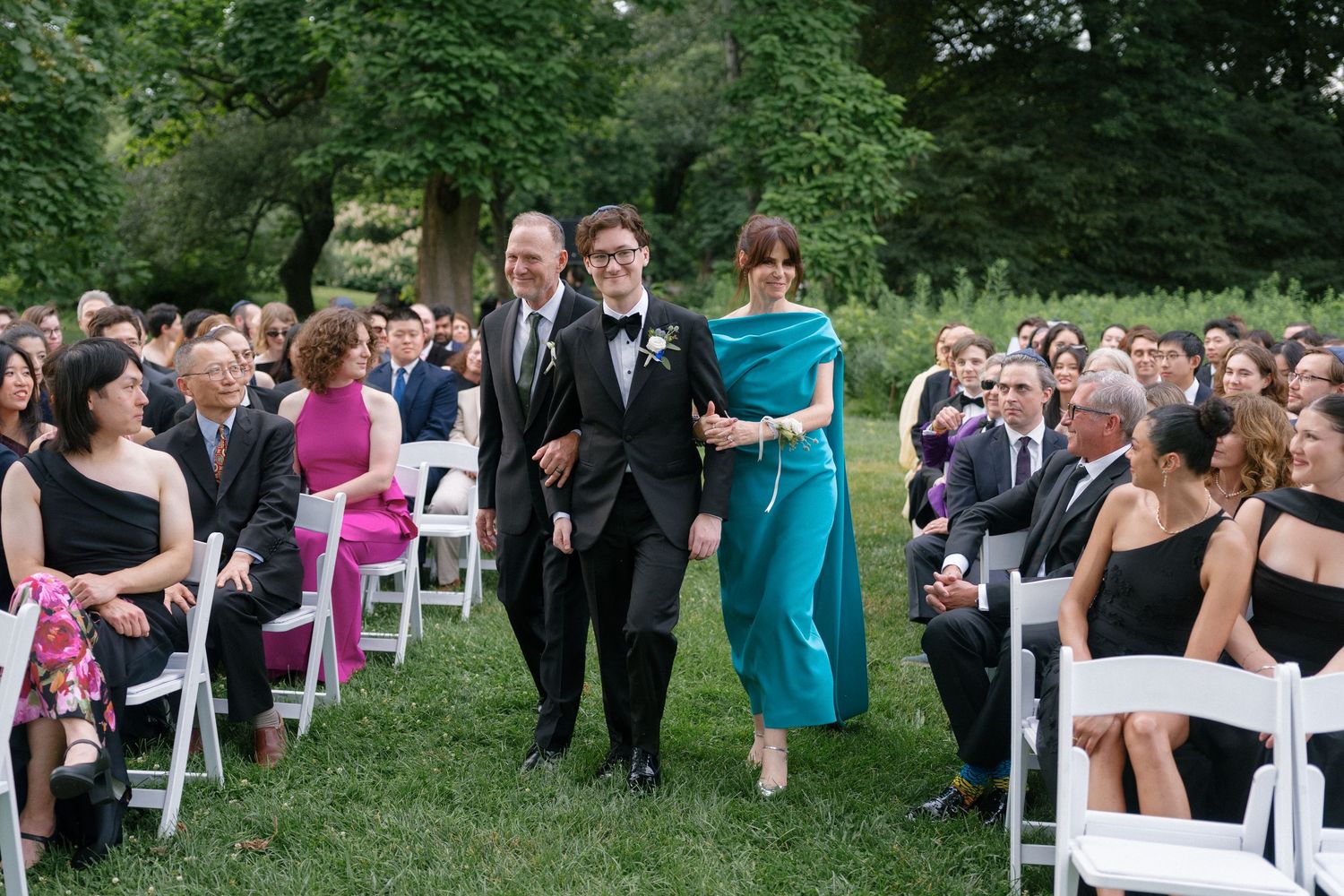 Groom walks down outdoor wedding aisle with parents, guests seated in white chairs on lush green lawn.
