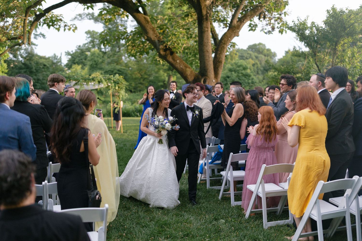 Bride and groom walk down outdoor wedding aisle surrounded by guests under lush green trees at garden ceremony.