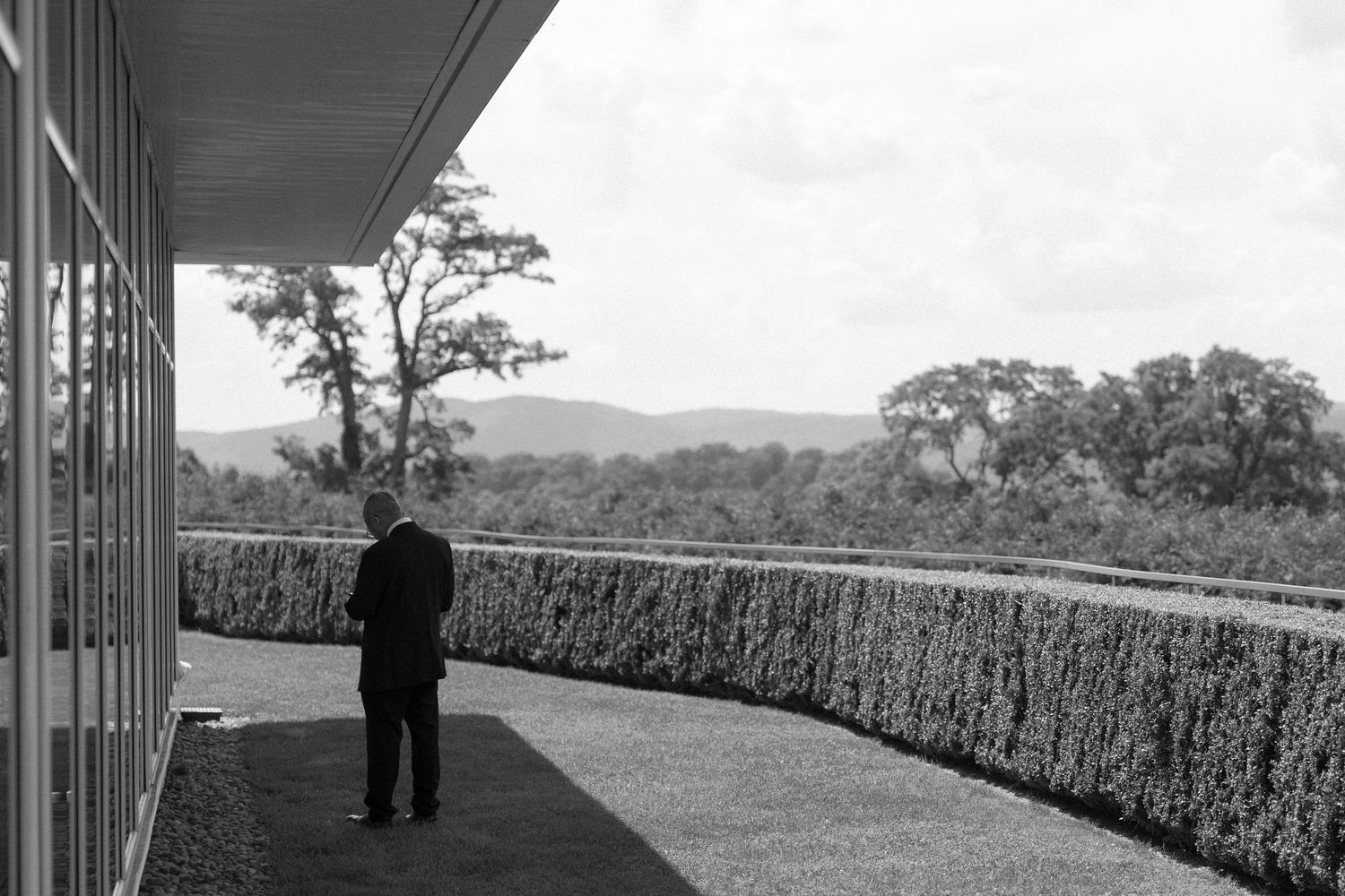 Black and white photo of a man standing on a curved terrace overlooking lush hedges and rolling hills.