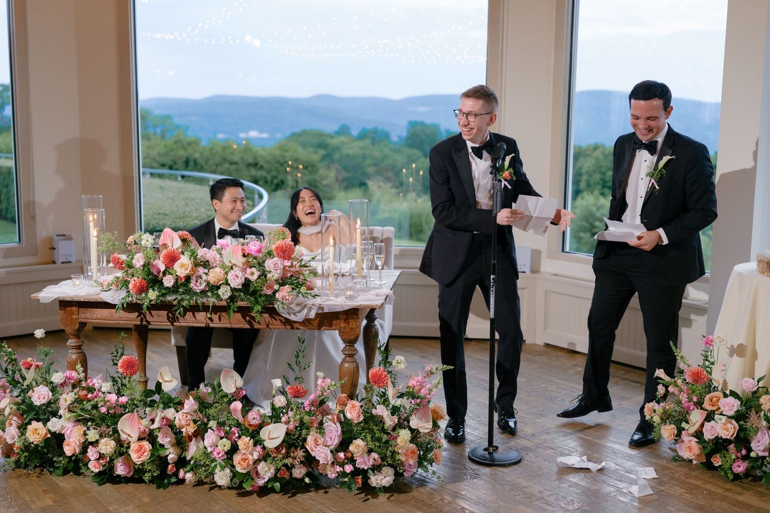 Two groomsmen delivering speech at sweetheart table adorned with lush pink floral arrangements and mountain backdrop.