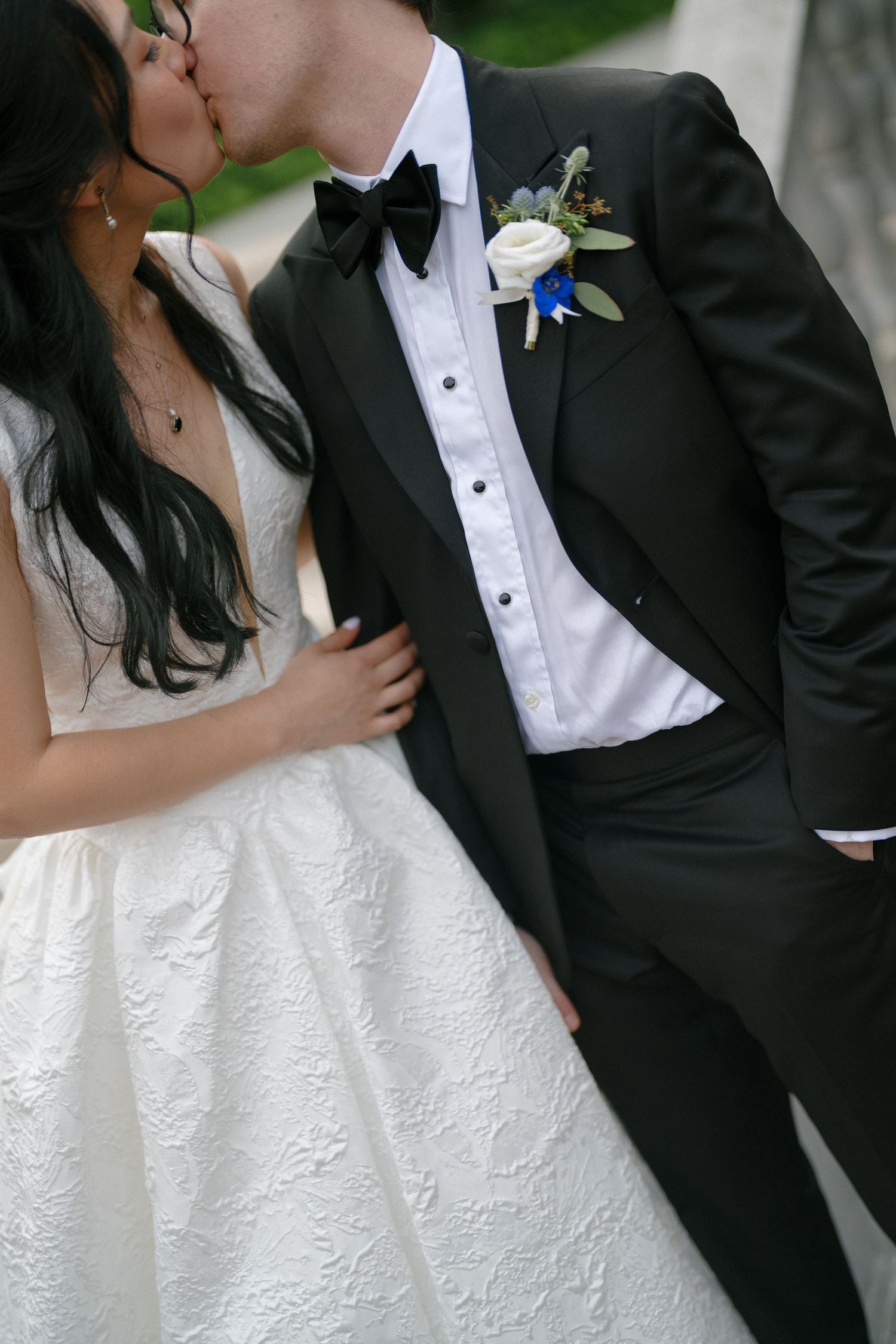 Newlyweds share a romantic kiss, groom in black tuxedo with boutonniere embraces bride in white lace gown.