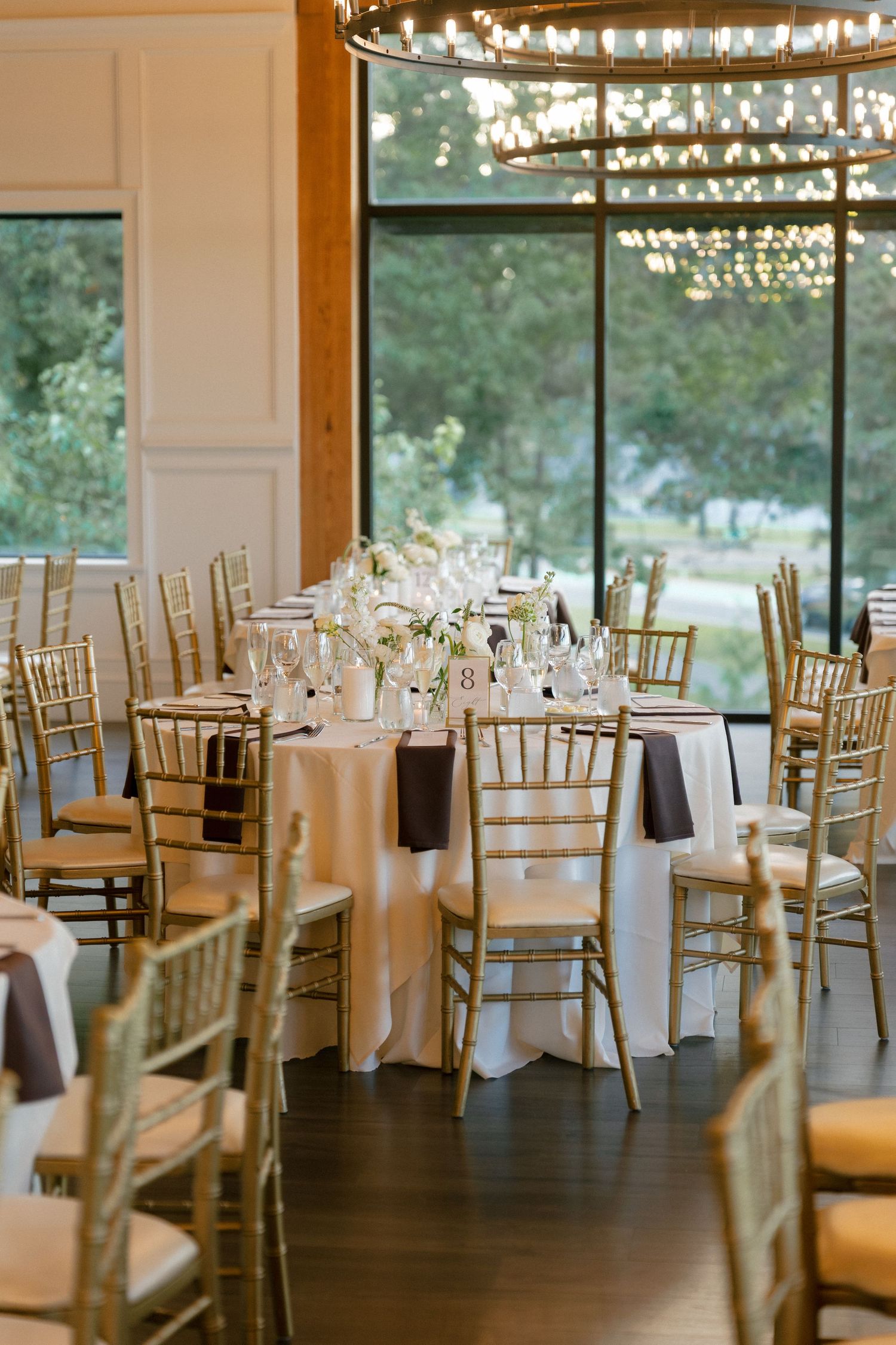 Elegant wedding reception hall with gold chiavari chairs, white tablecloths, floral centerpieces, and warm chandelier lighting.