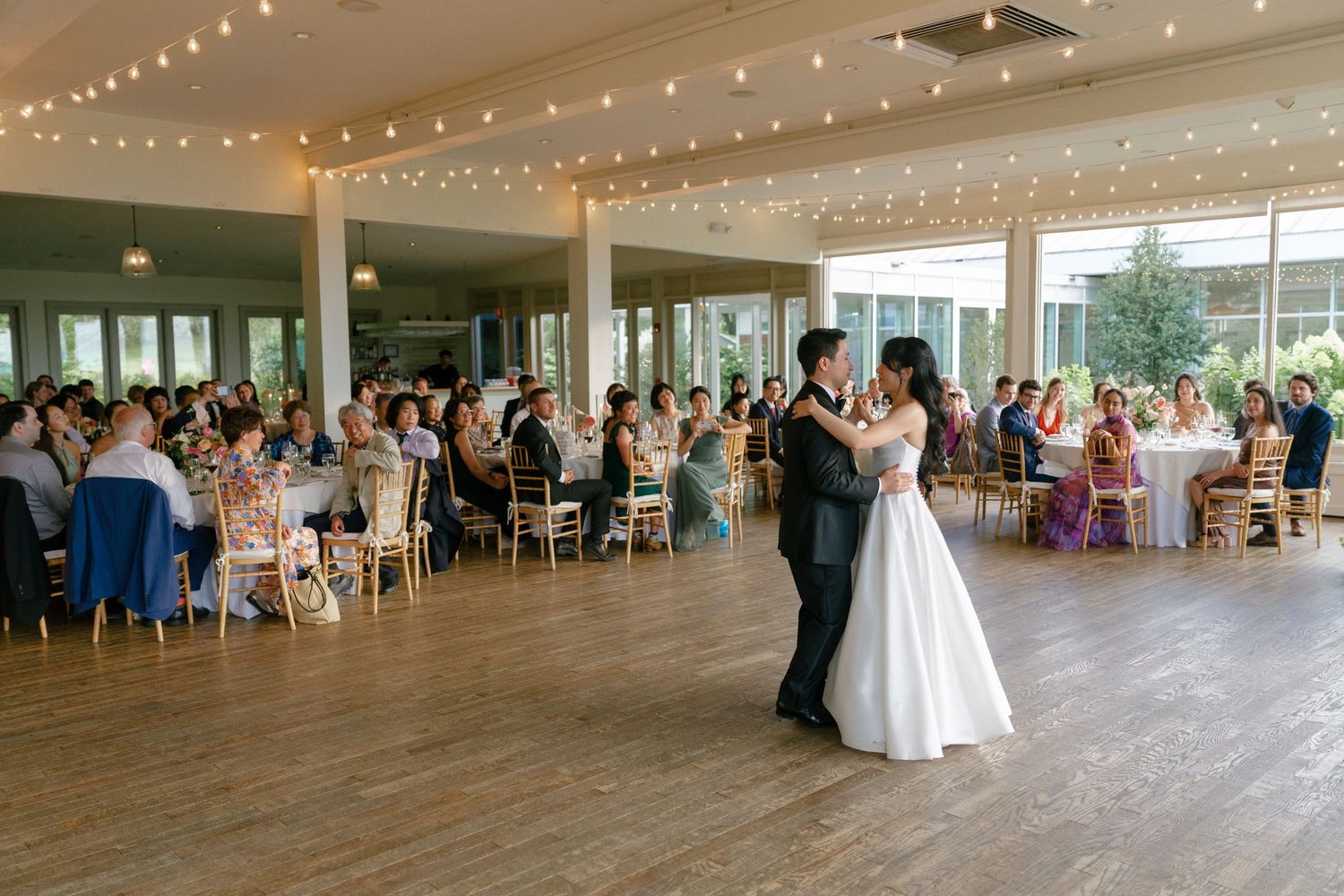 Bride and groom share first dance in elegant reception hall with string lights and seated wedding guests.