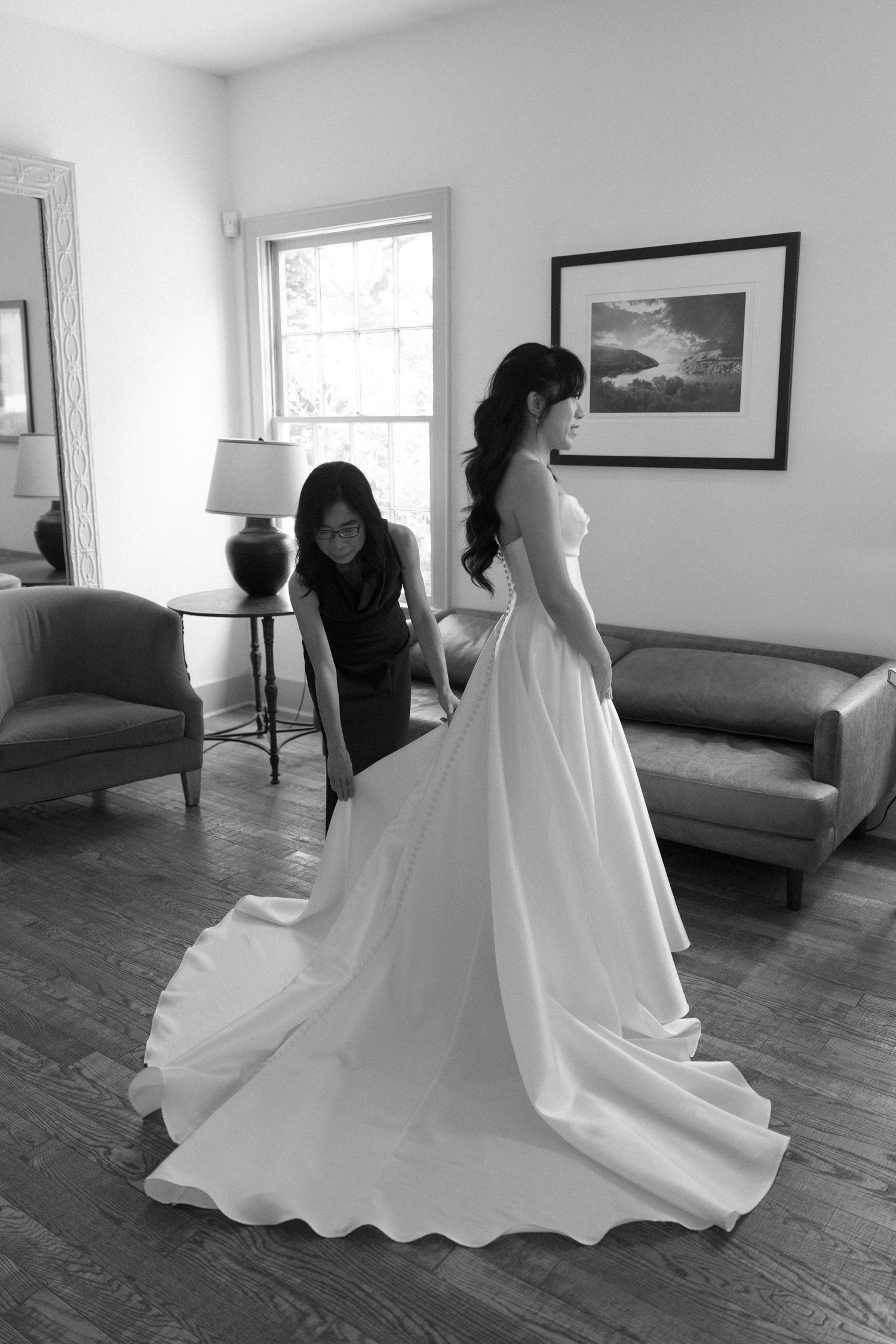 Black and white photo of a bride in a elegant gown being assisted by a bridesmaid in a cozy room.