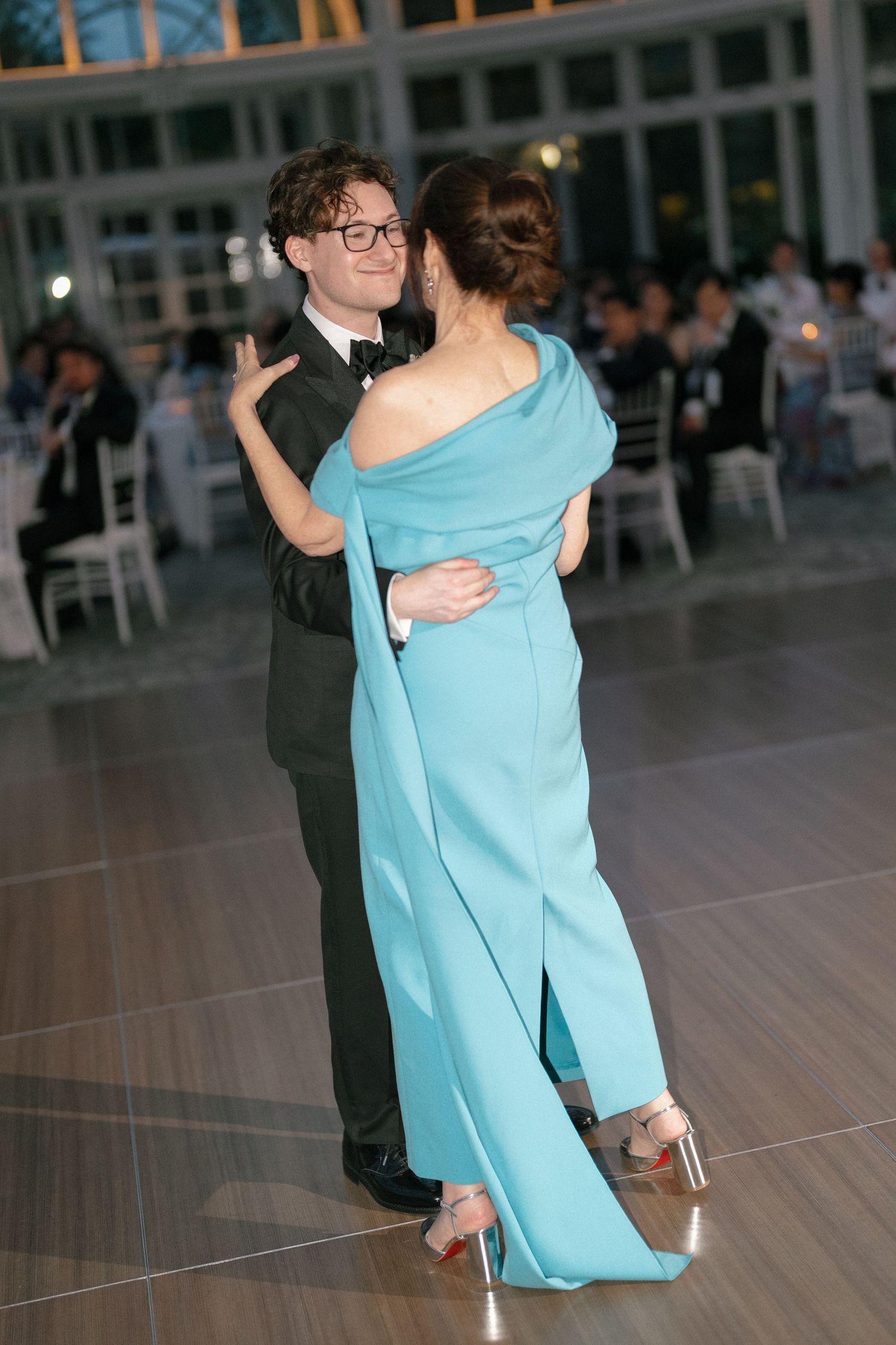 Young man in tuxedo shares mother-son dance with woman in elegant off-shoulder teal gown at reception.