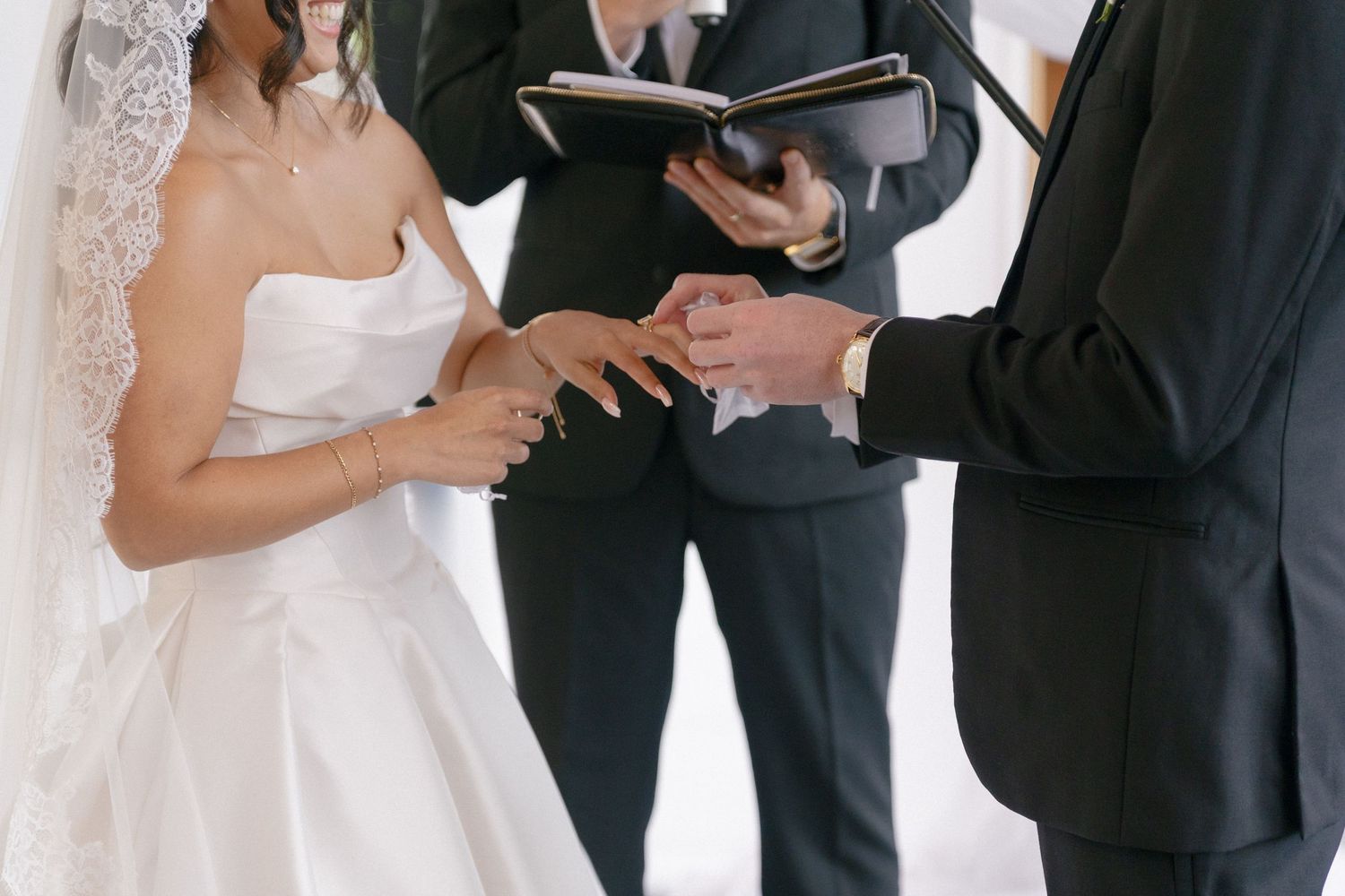Bride and groom exchanging rings during wedding ceremony with officiant holding book in background.