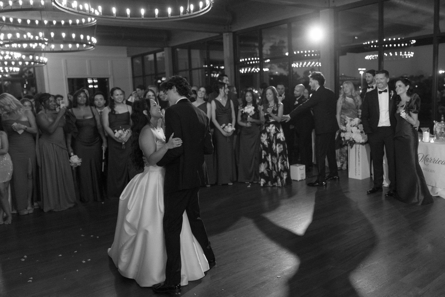 Bride and groom share first dance surrounded by guests in elegant black and white photo.