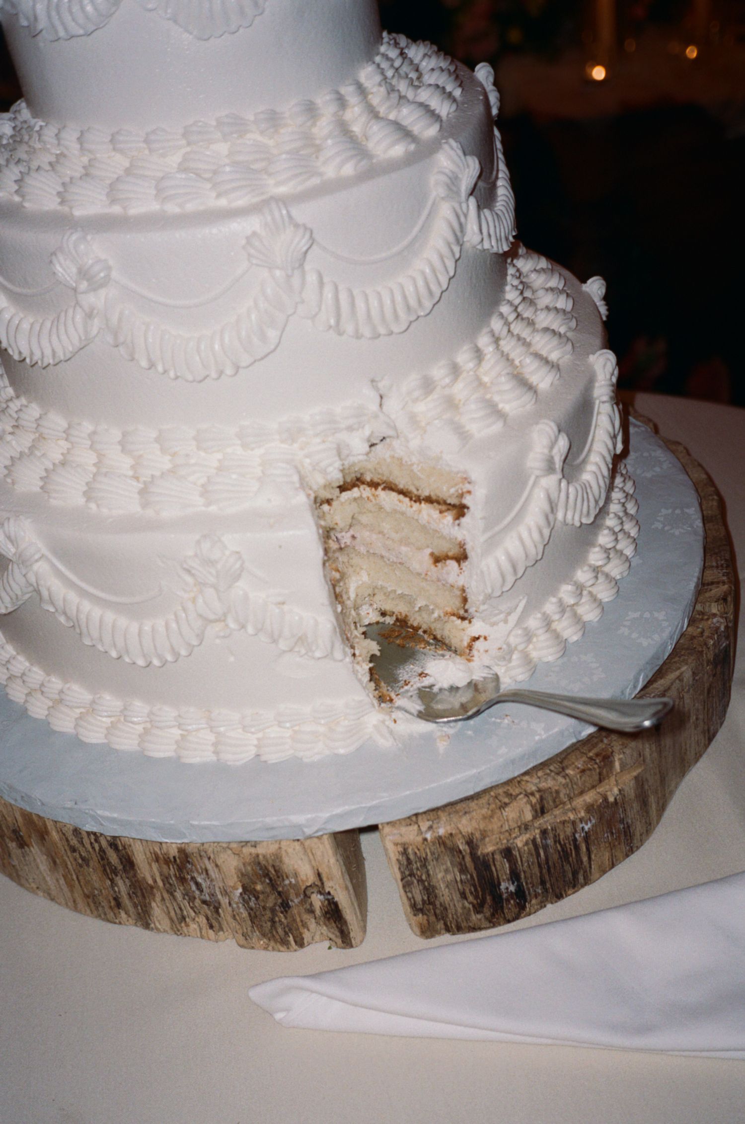 Elegant three-tier white wedding cake with decorative frosting on rustic wood slice stand, with a slice cut out revealing layers.