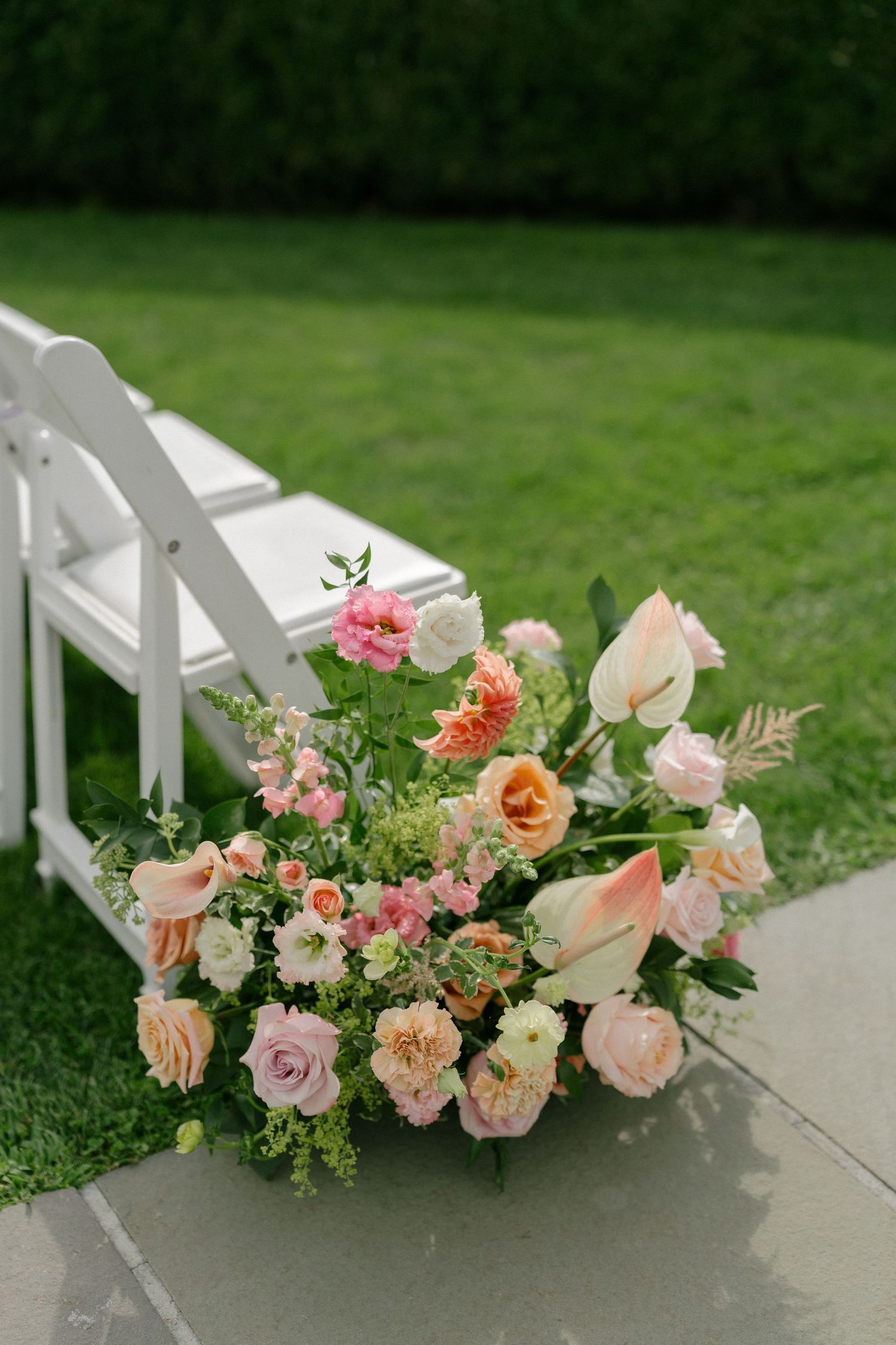 Lush peach, pink, and white floral arrangement with roses and tulips adorning white ceremony chair aisle.
