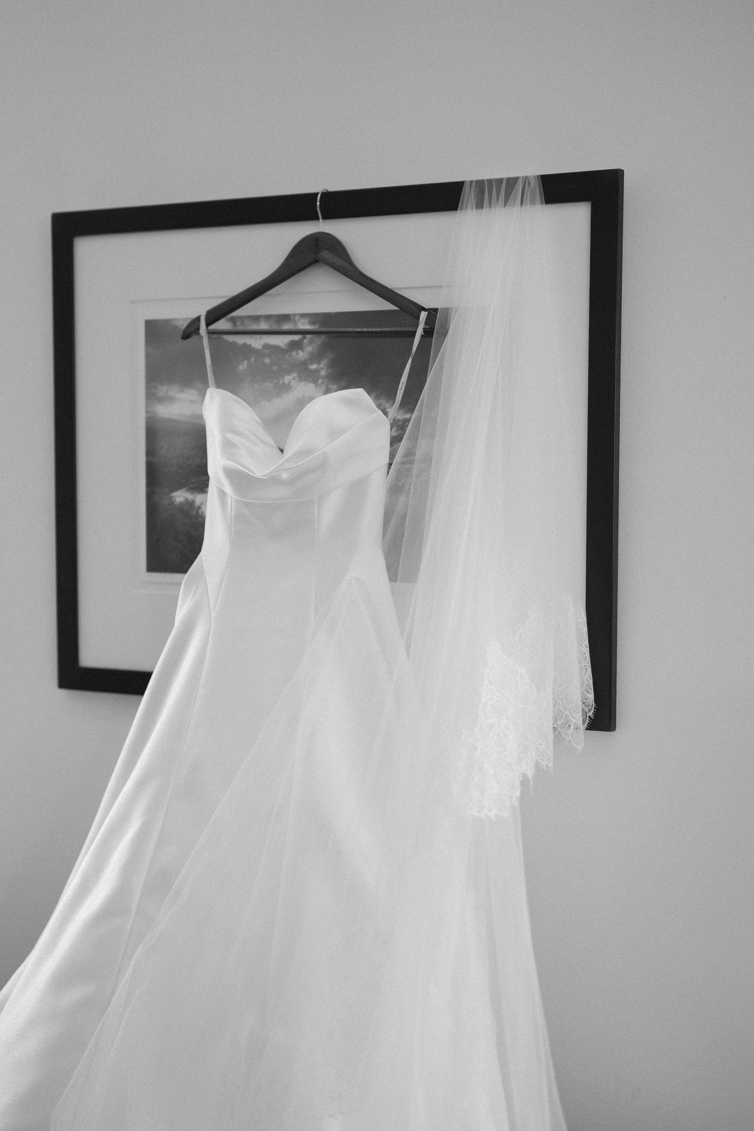 Black and white photo of a strapless wedding gown with veil hanging on a wooden hanger.