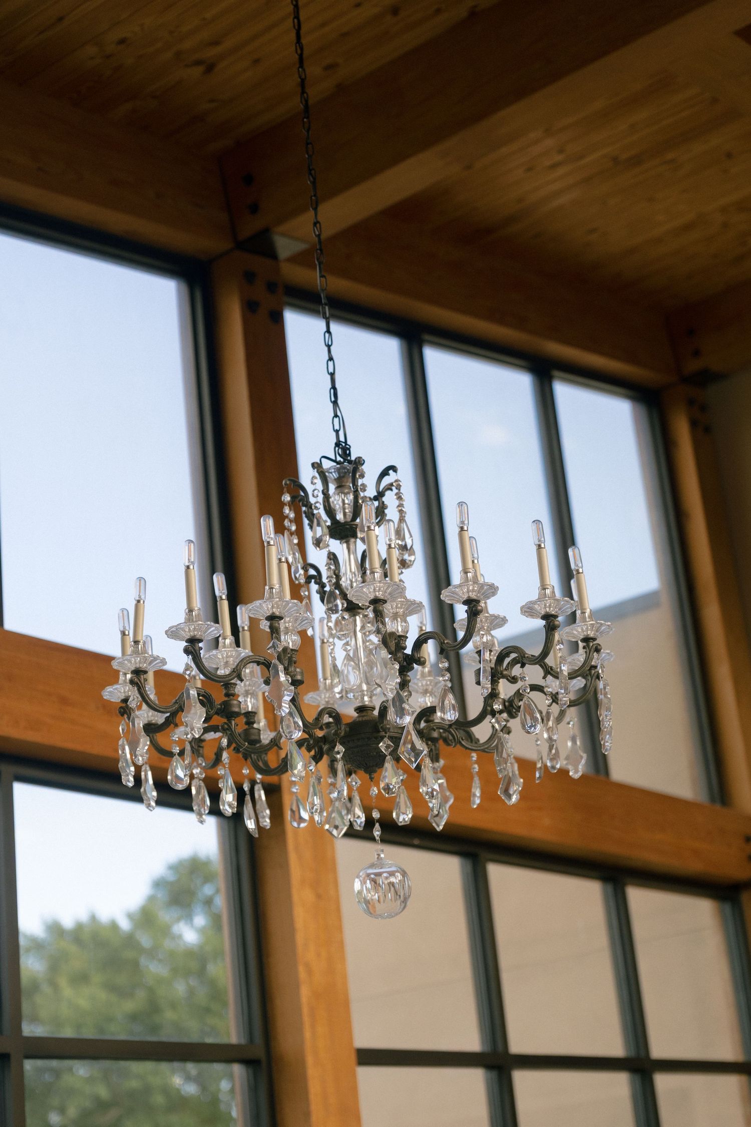 Ornate crystal and iron chandelier hanging in rustic wooden venue with large windows and blue sky backdrop.