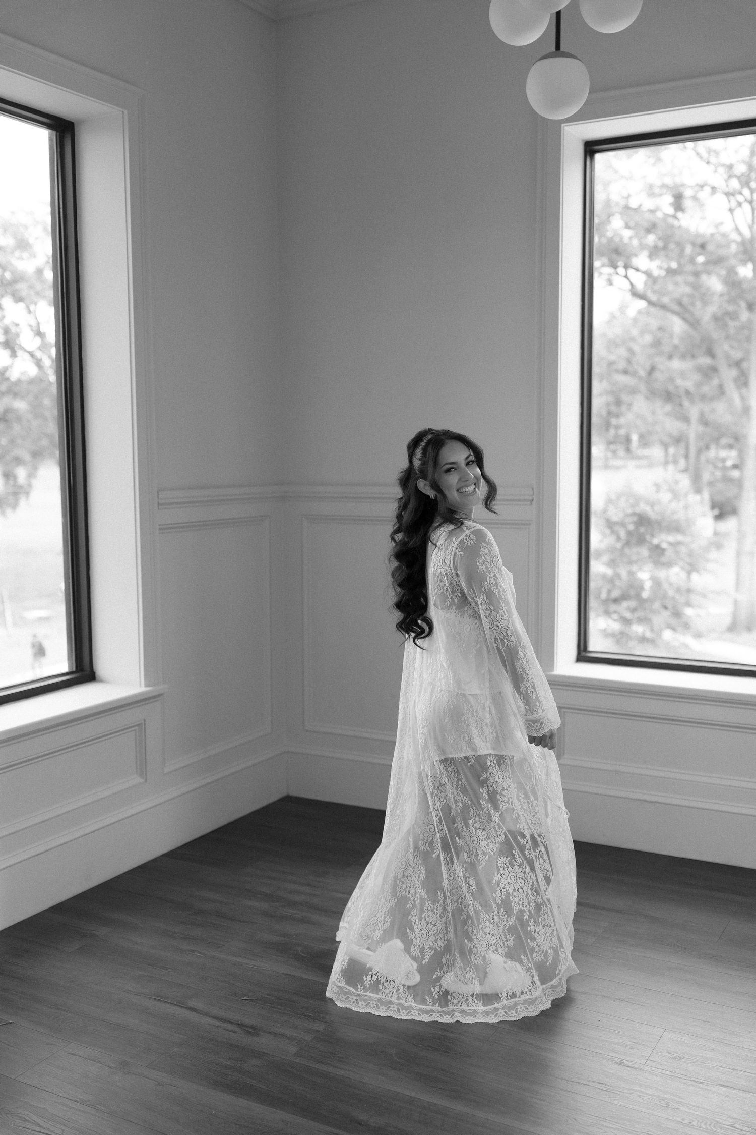 Bride in lace robe smiling over shoulder in black and white photo by sunlit windows.