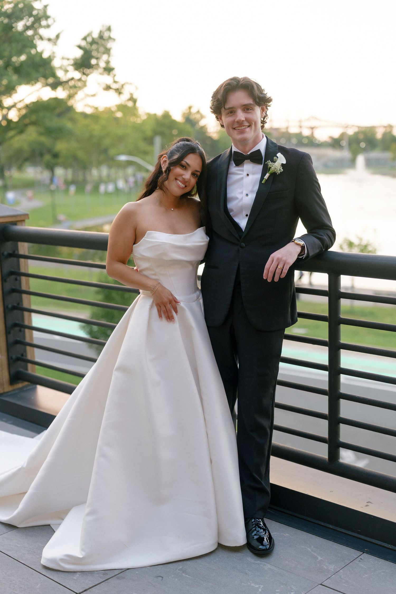 Bride in strapless white ballgown and groom in black tuxedo pose together on outdoor terrace at sunset wedding.