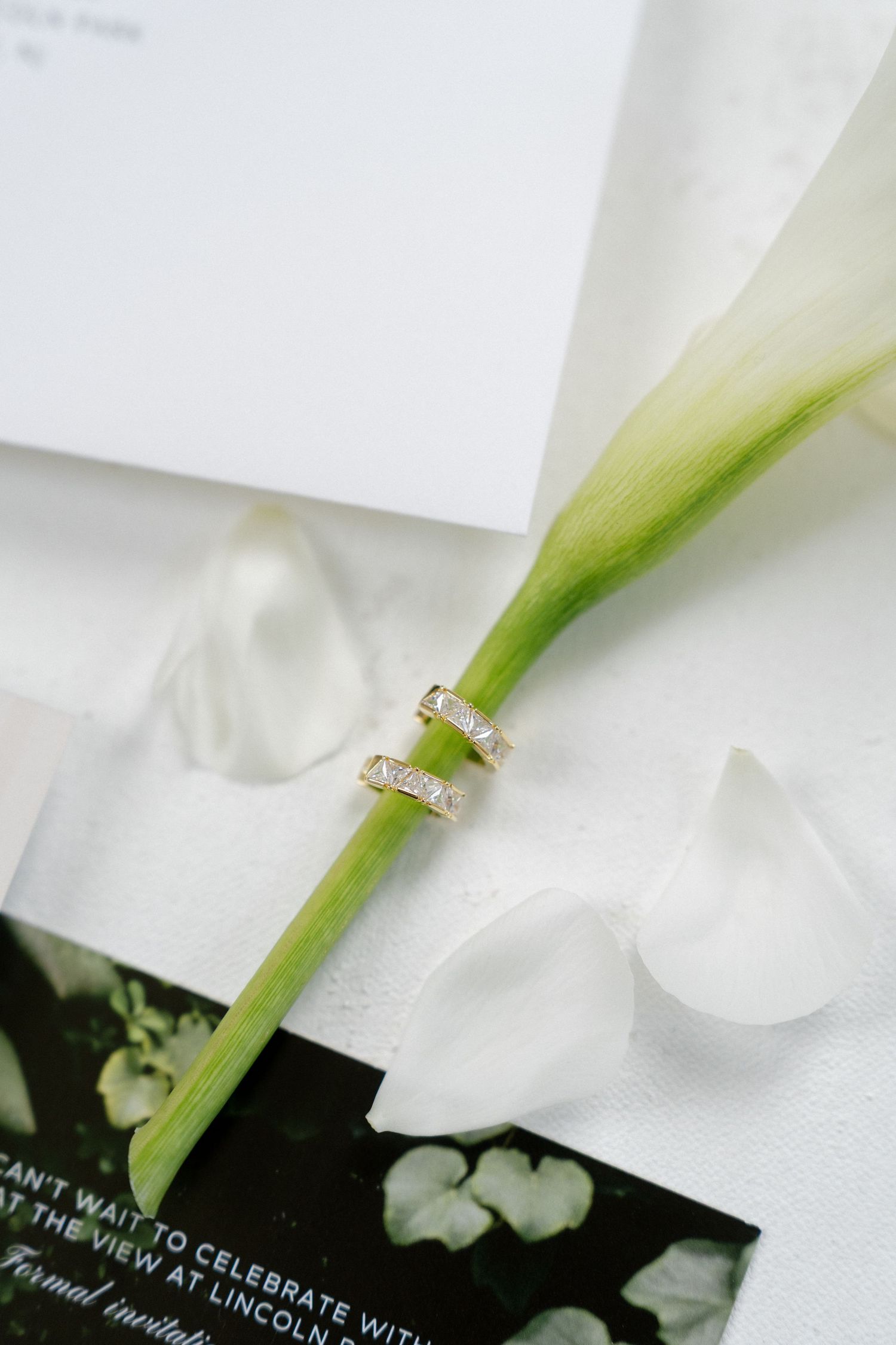 Gold wedding bands resting on a white calla lily stem with wedding invitation.