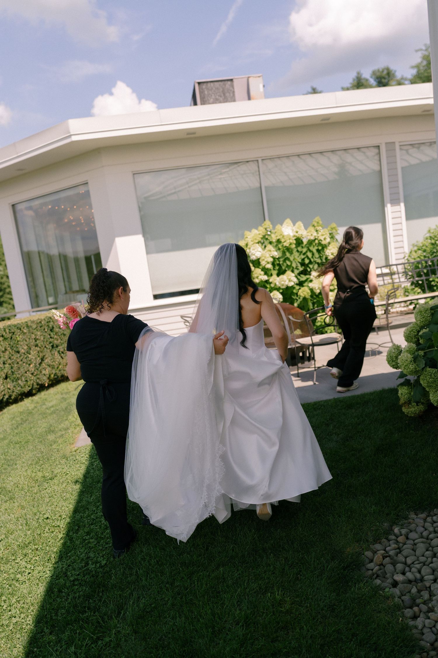 Wedding attendants helping bride with white veil and train walking across green lawn outdoors.