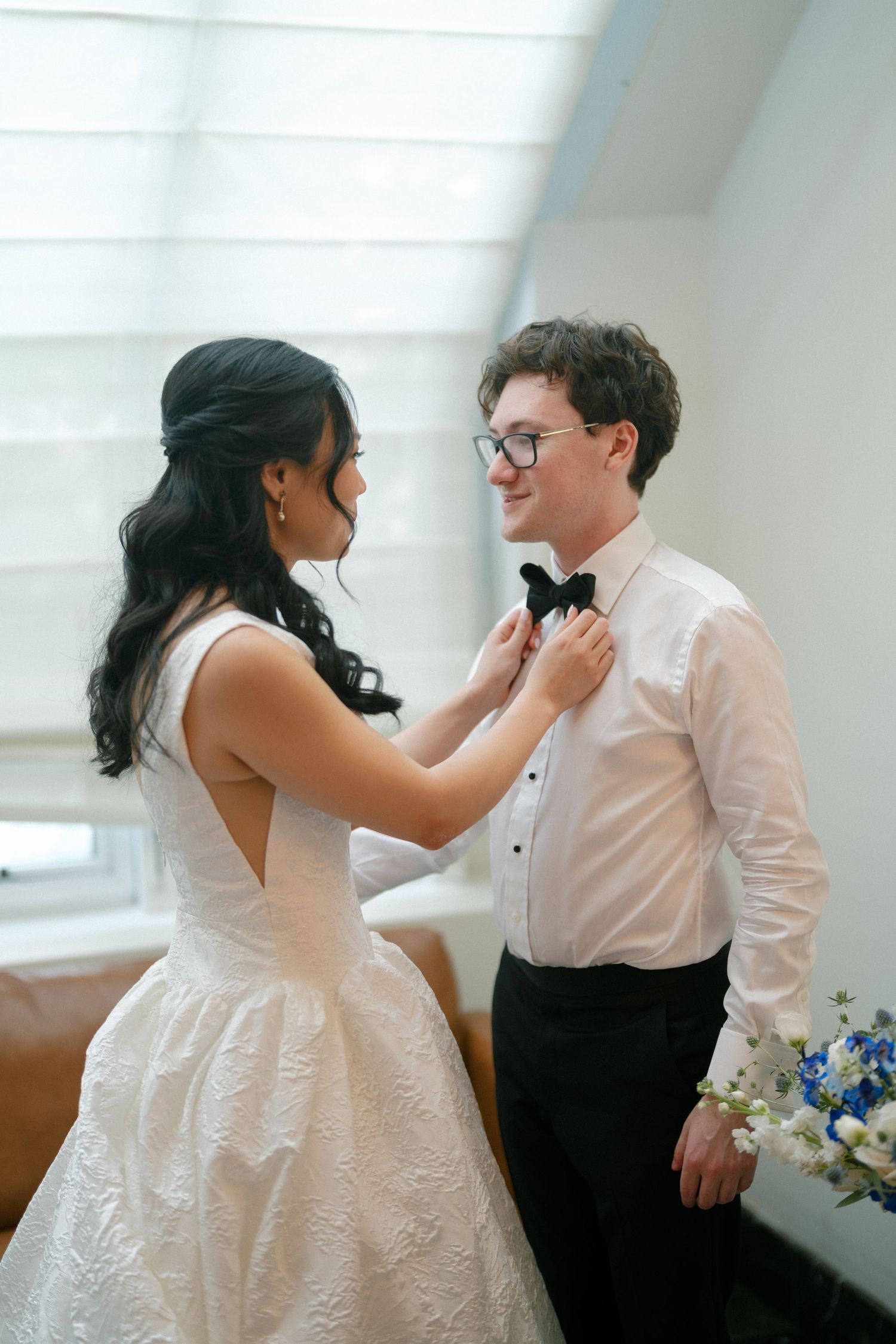 Bride in white dress adjusting bow tie of smiling groom wearing glasses in bright indoor setting.