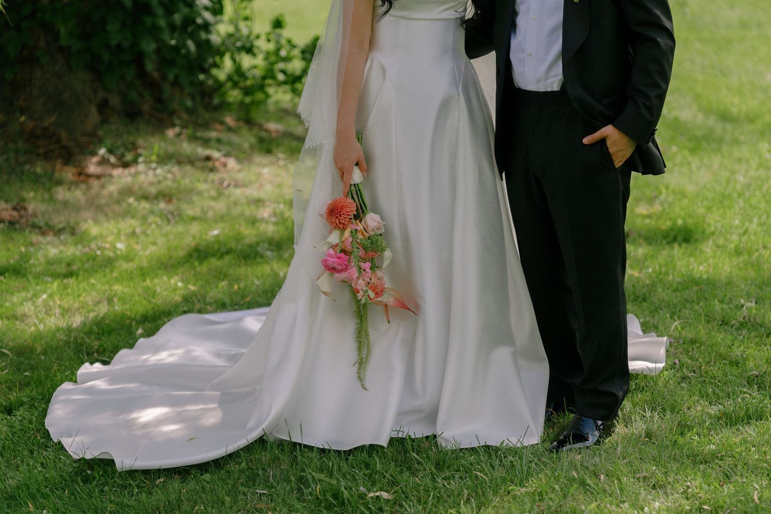 Bride in white gown holding pink bouquet stands beside groom in black tuxedo on green grass.