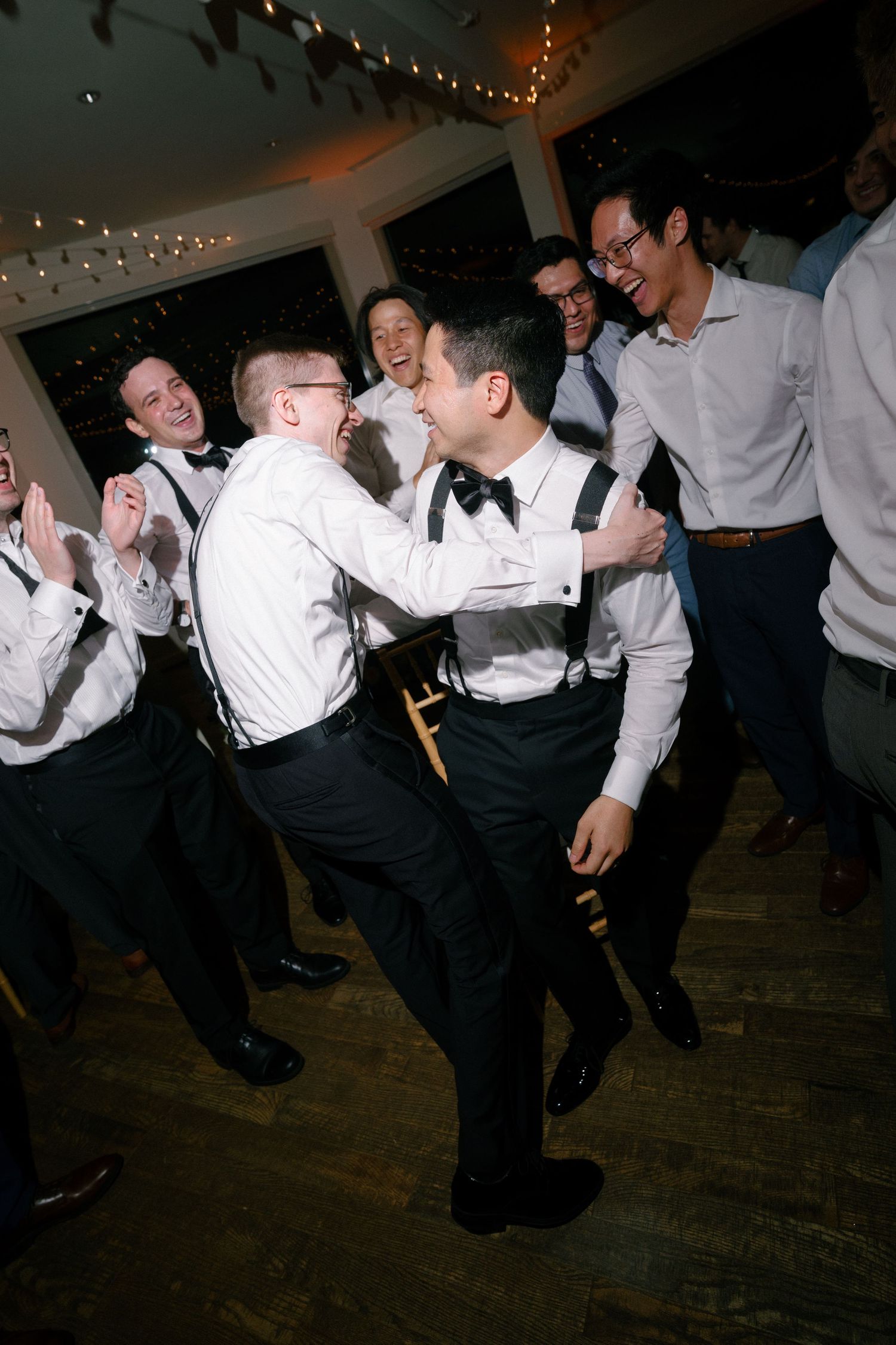 Groomsmen in black and white attire laugh and dance together at a lively indoor wedding reception celebration.