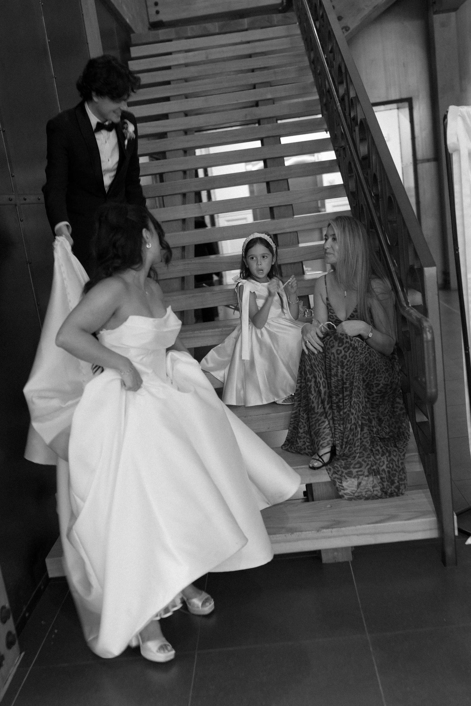 Black and white photo of bride, groom, flower girl, and guest gathered on wooden stairs at wedding.