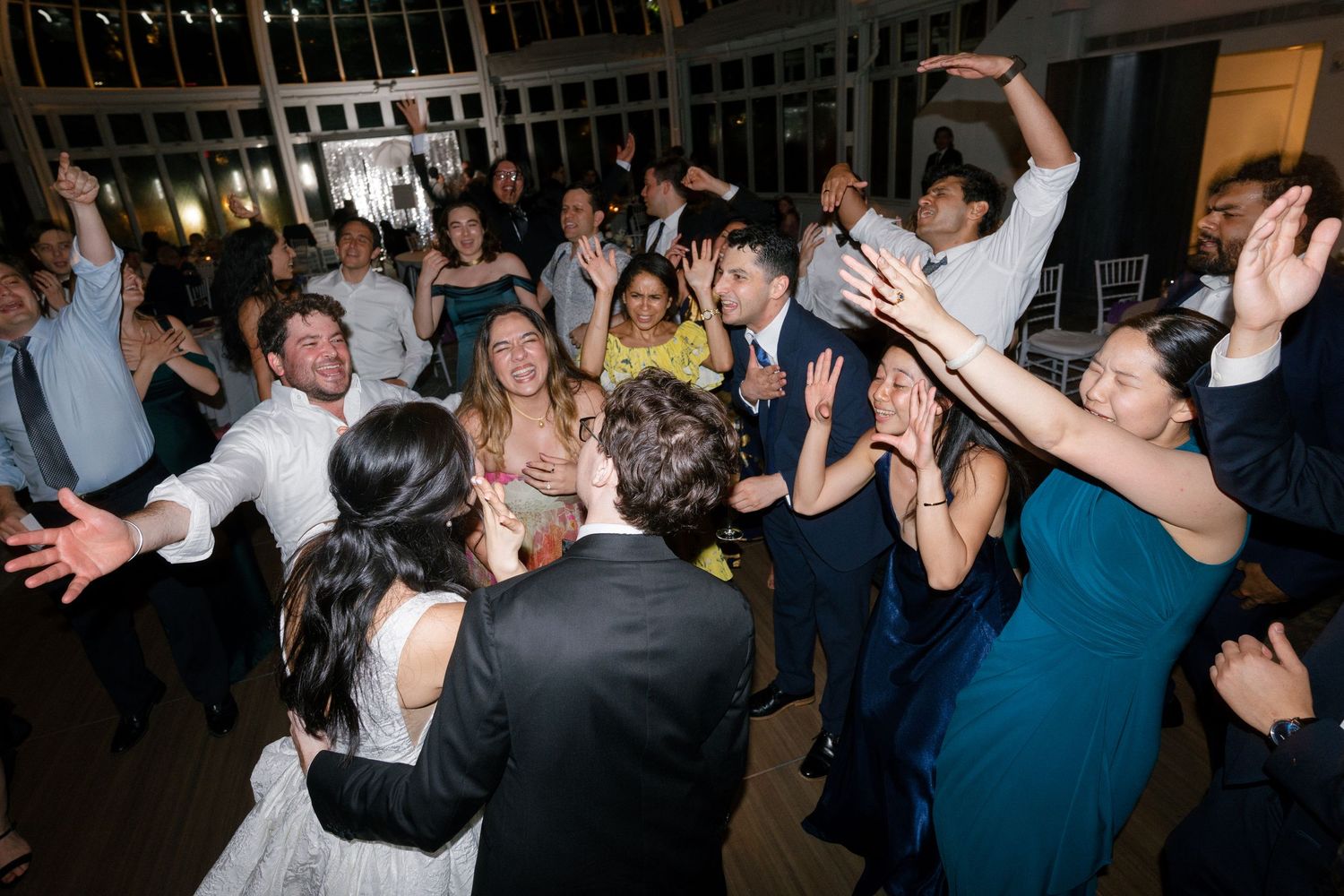Joyful wedding reception dance floor with bride and groom surrounded by cheering guests with raised arms.