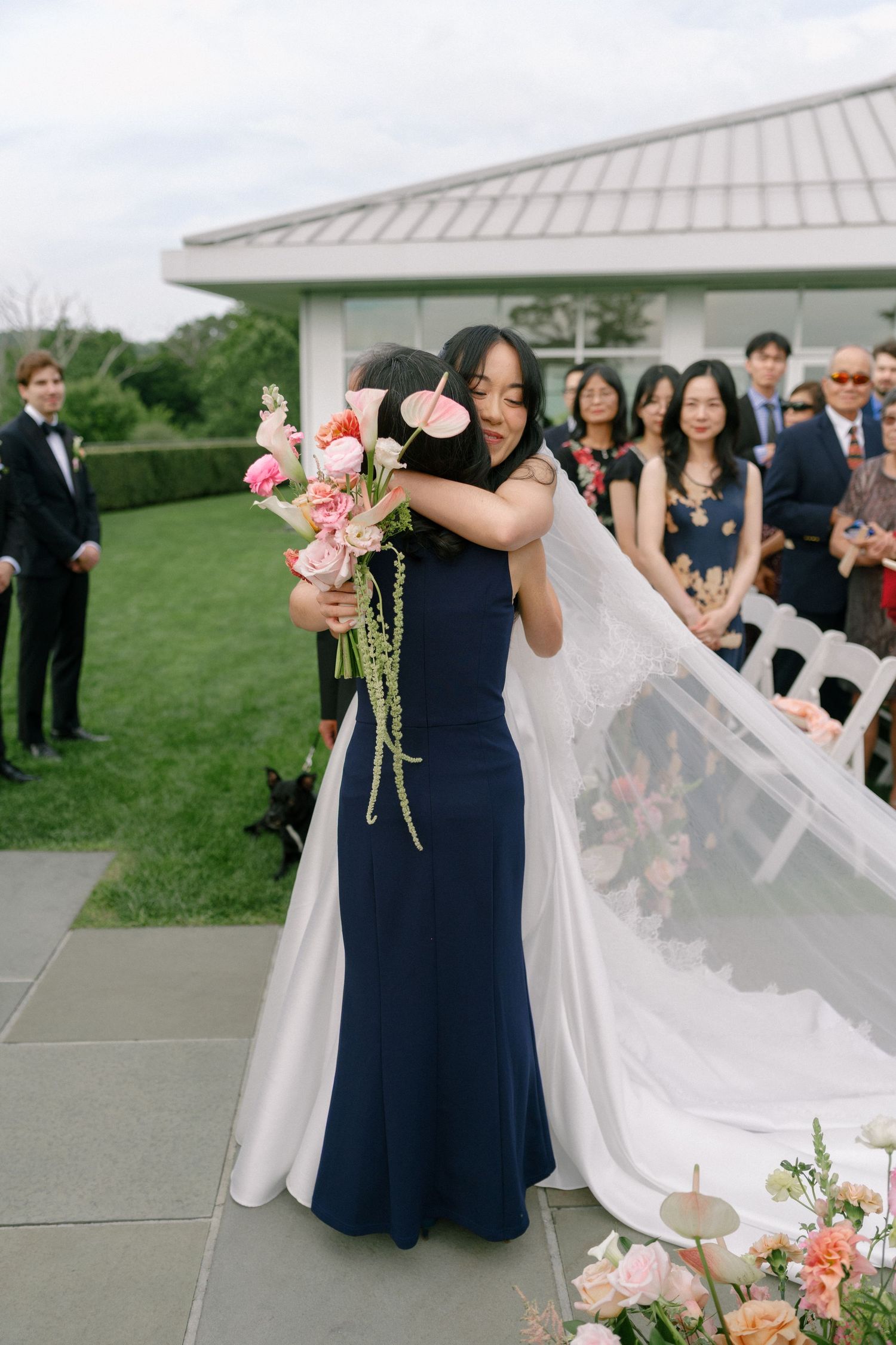Bride in white gown embracing bridesmaid in navy dress while holding pink floral bouquet at outdoor ceremony.