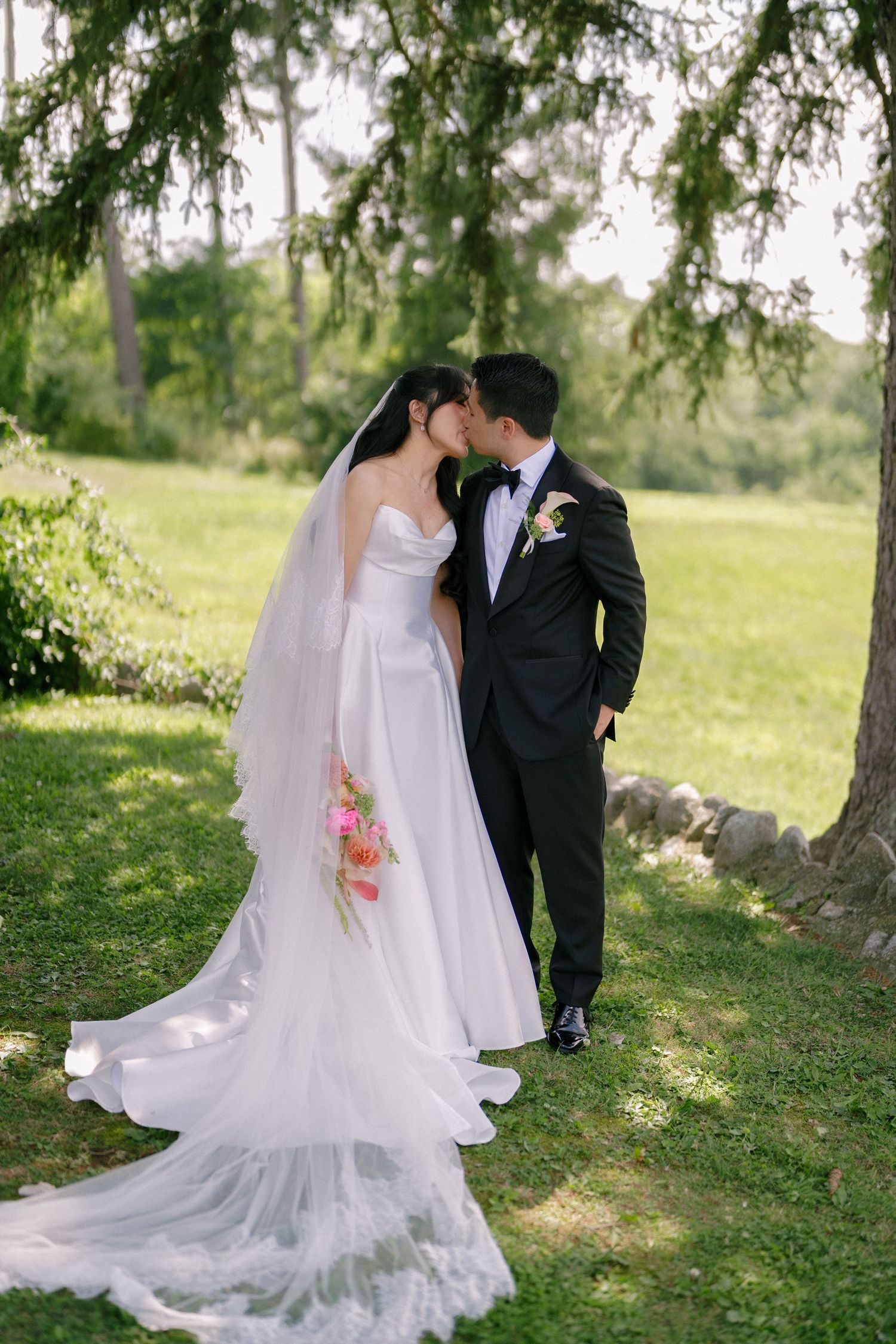 Bride and groom share romantic kiss under weeping tree in lush green garden after ceremony.