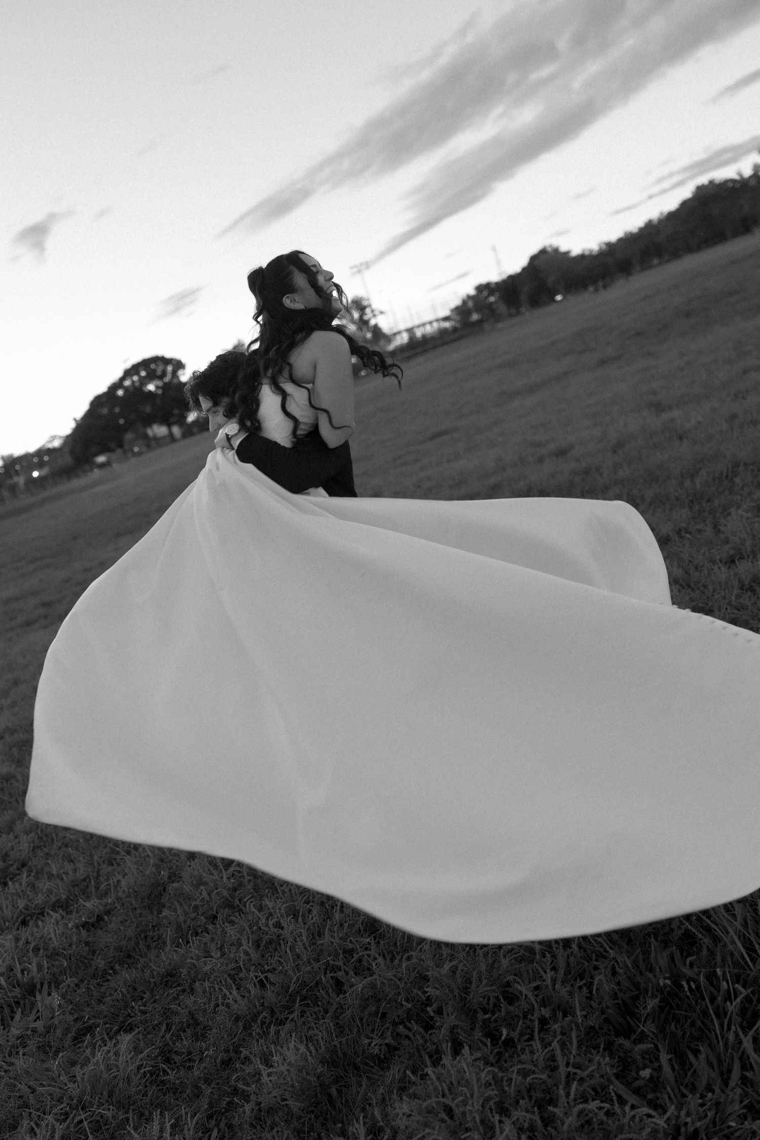 Black and white photo of a woman in a flowing gown with fabric billowing in the wind in an open field at dusk.