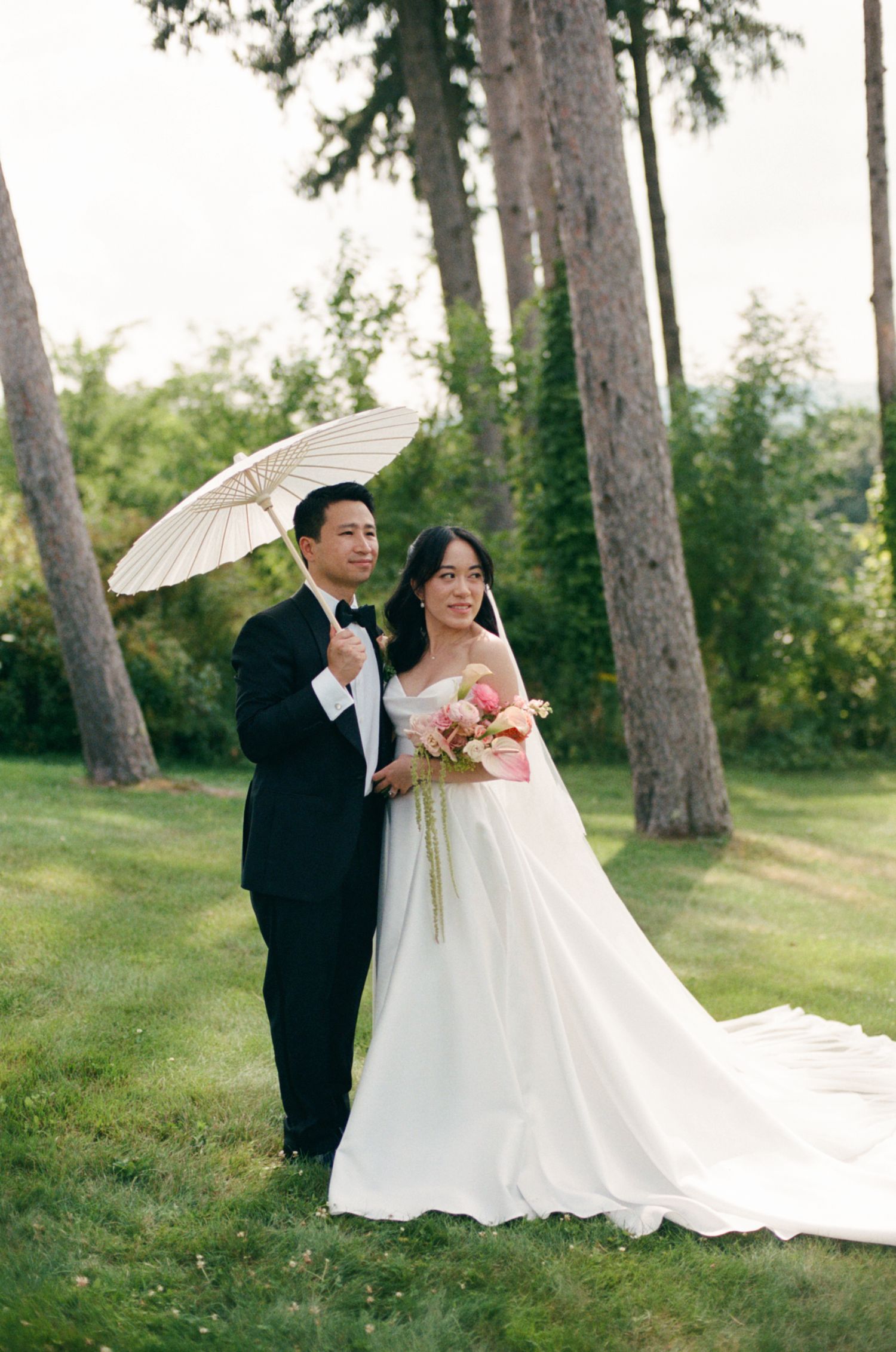 Bride and groom pose outdoors under a white parasol, bride holding pink bouquet in elegant white ball gown.