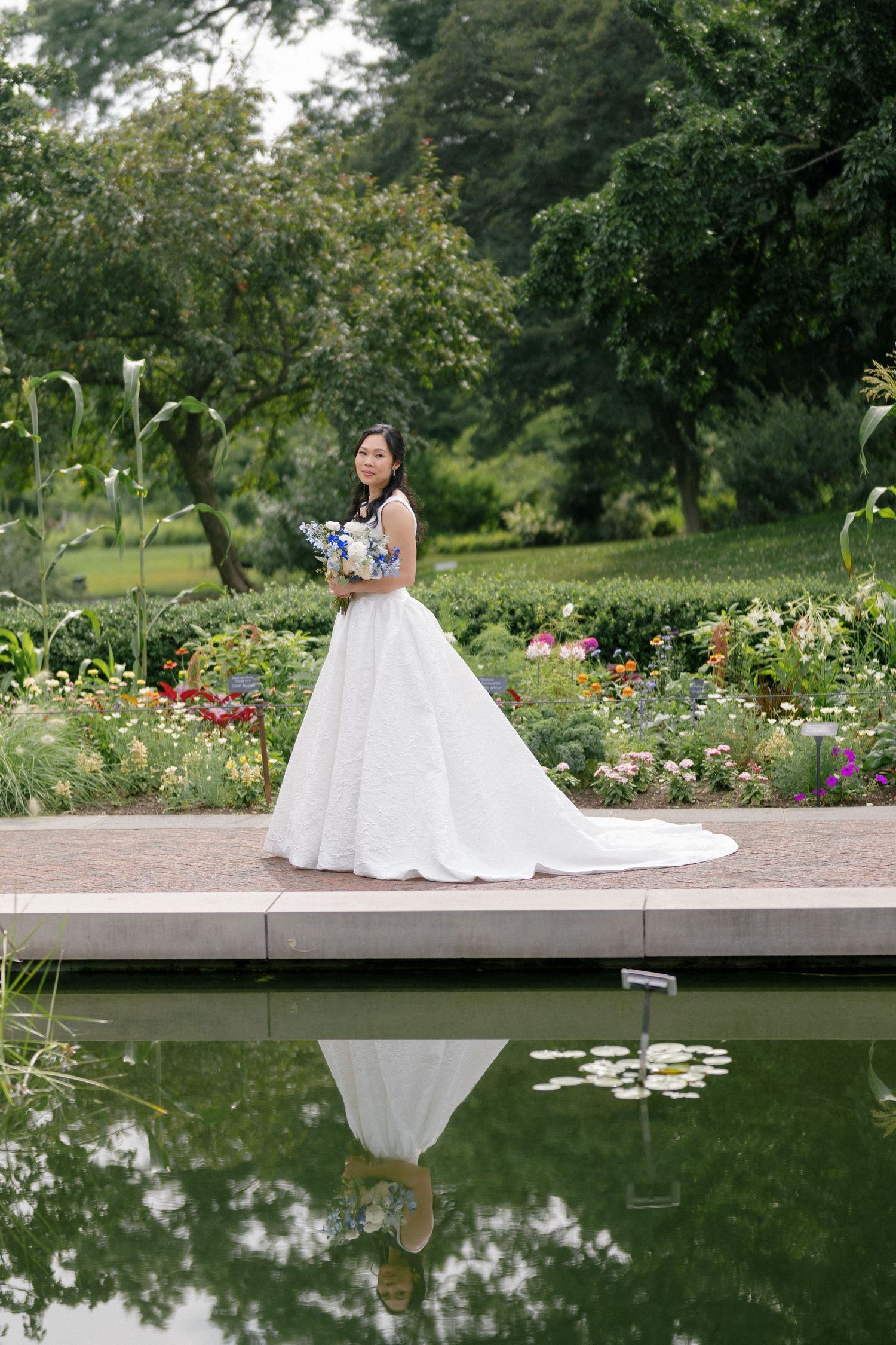 Bride in white ball gown holds bouquet beside garden pond, her reflection visible in the still water.