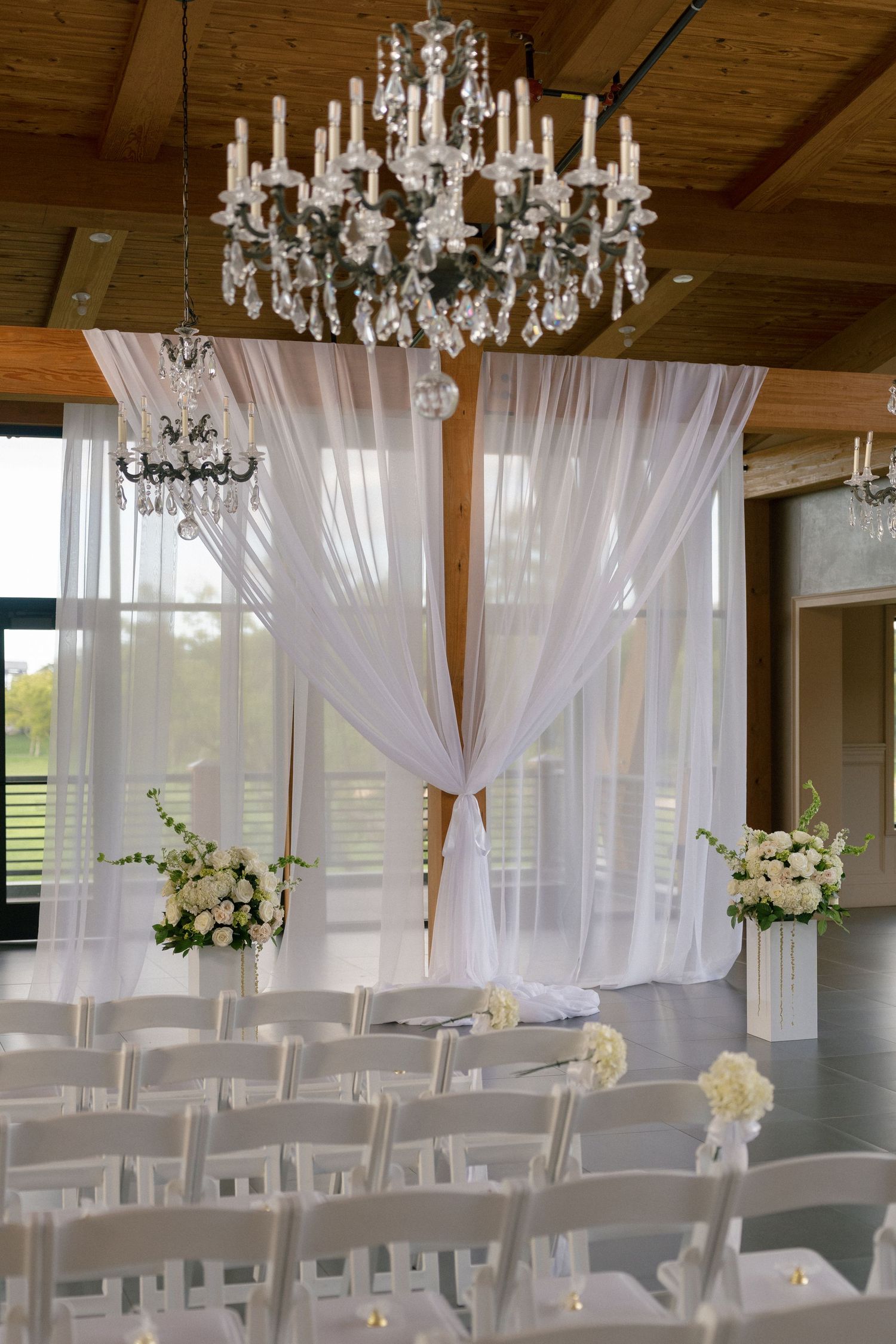 Elegant wedding ceremony venue with white draped altar, crystal chandeliers, and white chairs in rows.